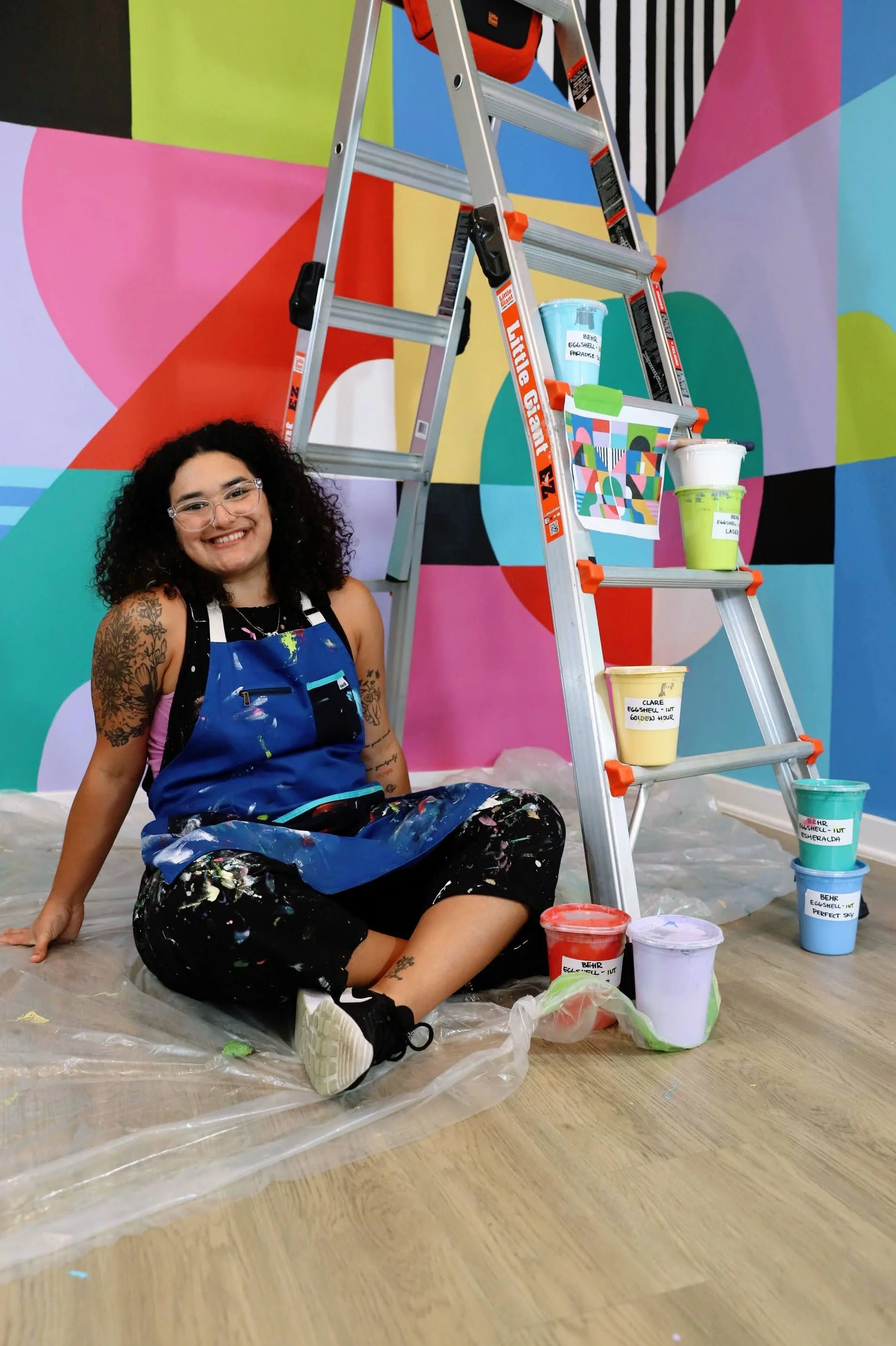 A woman sitting on the floor with a smile, surrounded by colorful paint buckets, in front of a vibrant, geometric mural. There is a ladder next to her, with some paint cans placed on its steps.