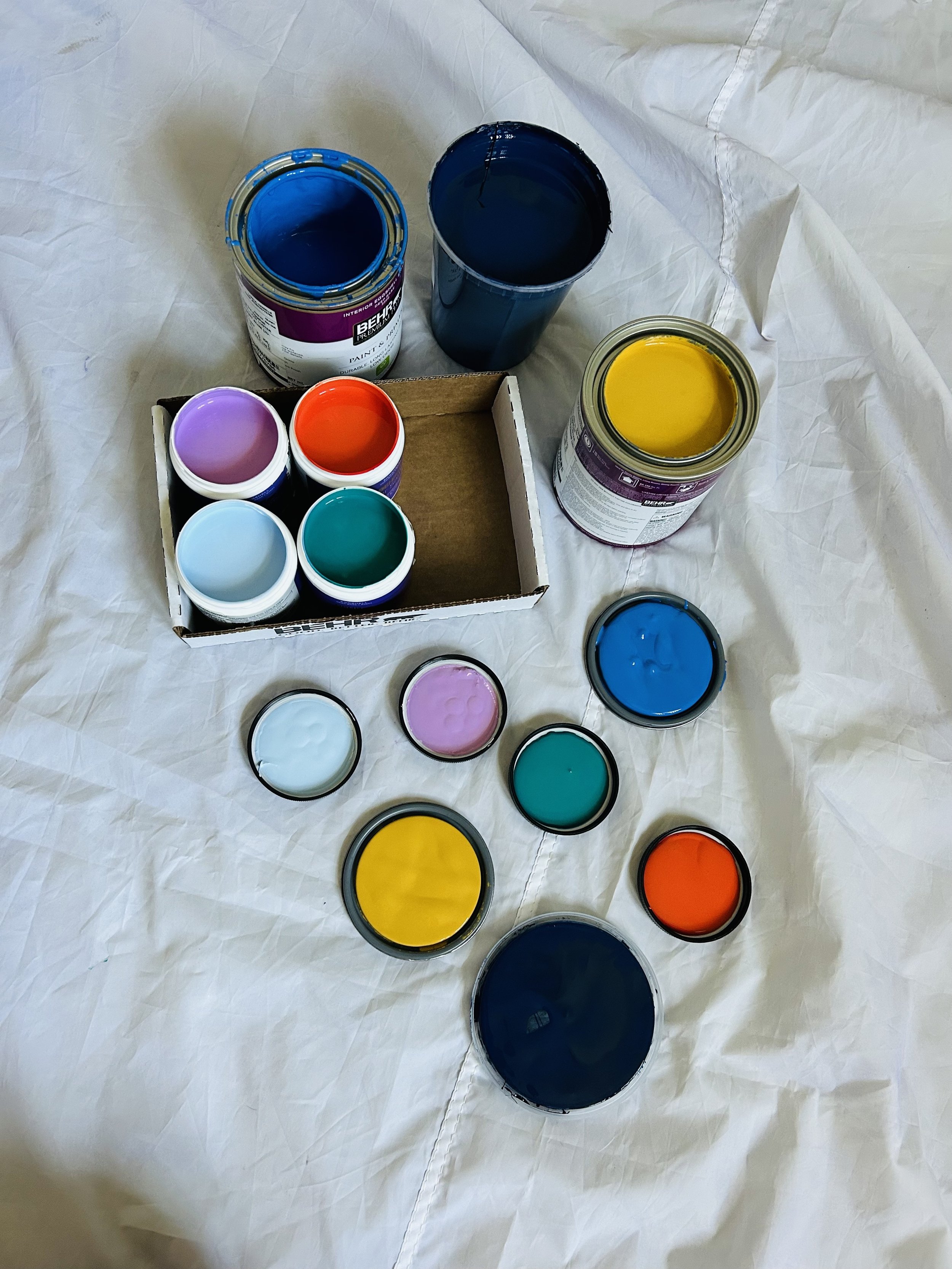 Open jars of blue, yellow, purple, teal, light blue, and orange paint, with some jars placed on a white cloth and a cardboard box.