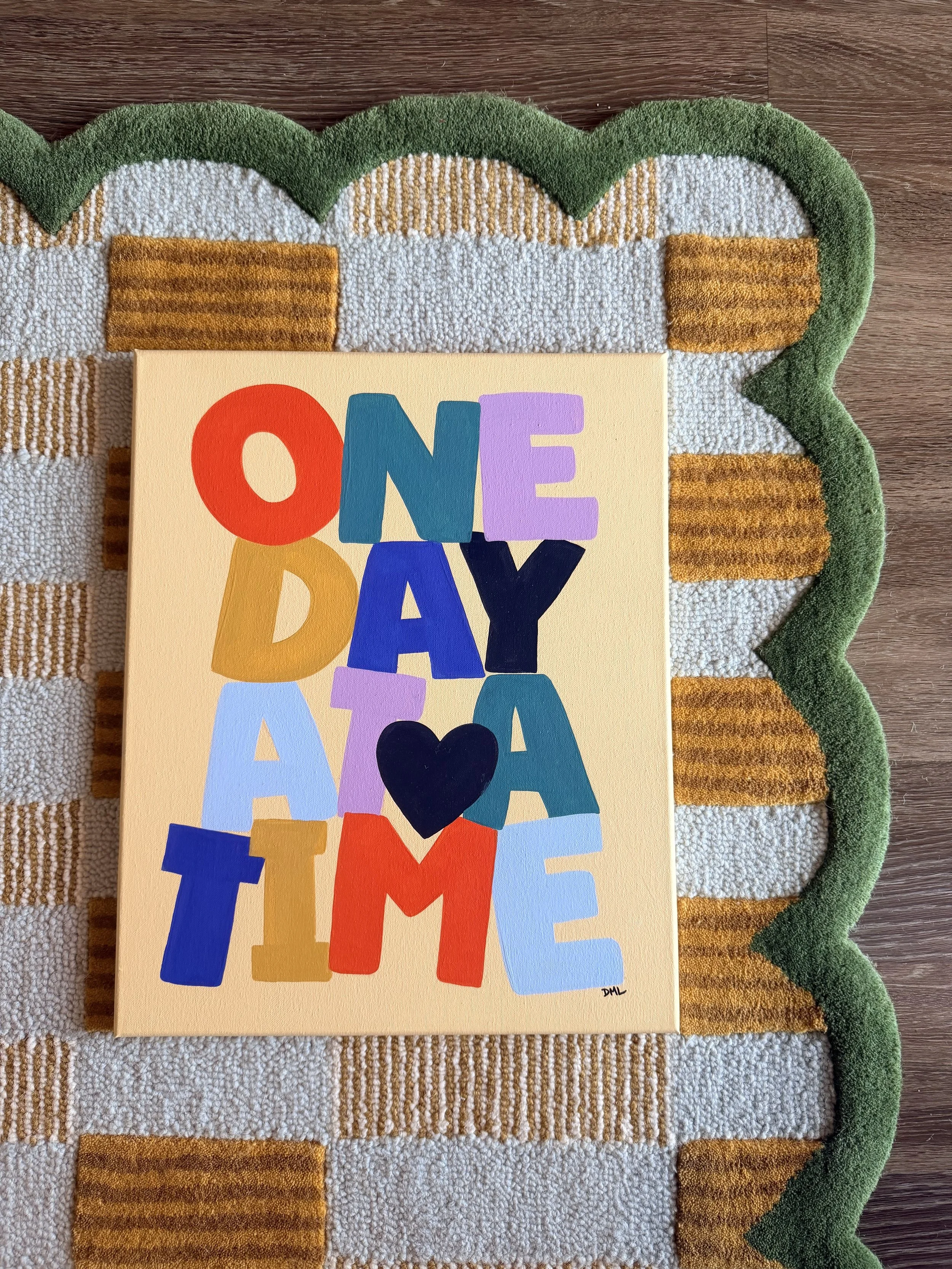 Colorful birthday card with the words "ONE DAY AT A TIME" and a black heart, placed on a striped green, white, and brown textured rug.