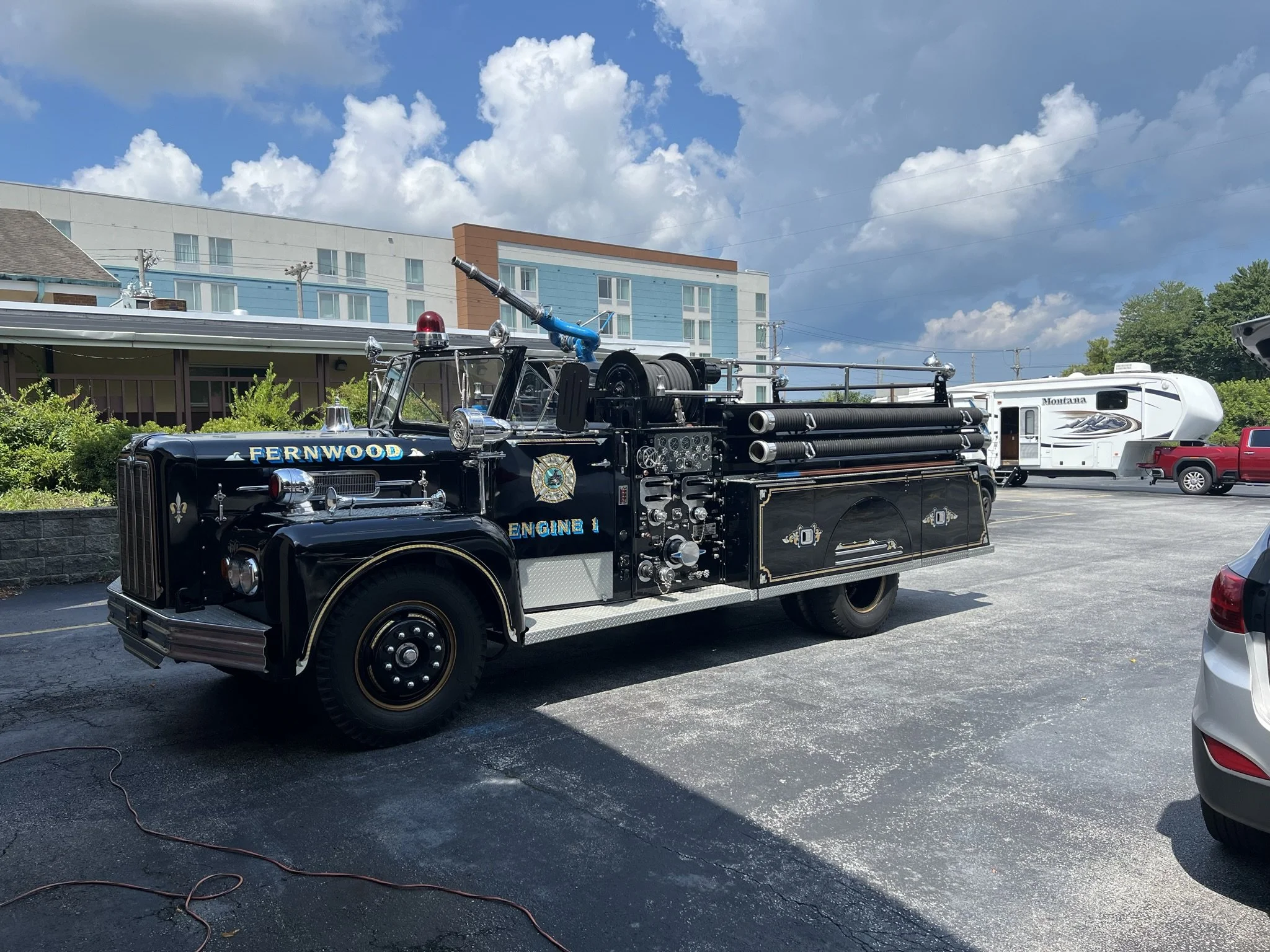 A vintage fire truck labeled 'Fernwood Engine 1' parked on an asphalt lot, with a white RV and red pickup truck in the background under a partly cloudy sky.