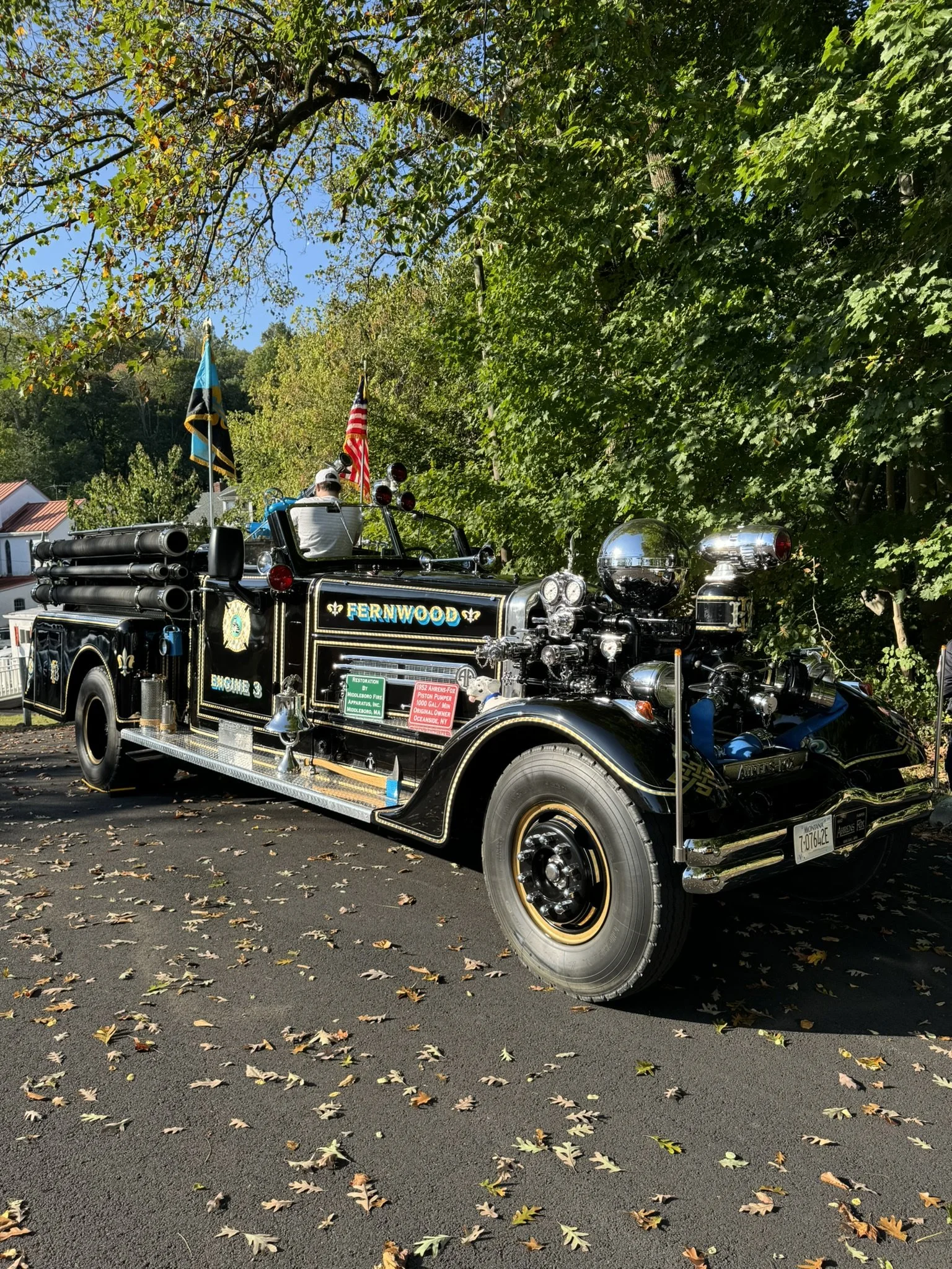 A vintage black fire truck with gold accents labeled "Fernwood" parked under green trees, with flags, hoses, and equipment on the truck, and fallen autumn leaves on the ground.