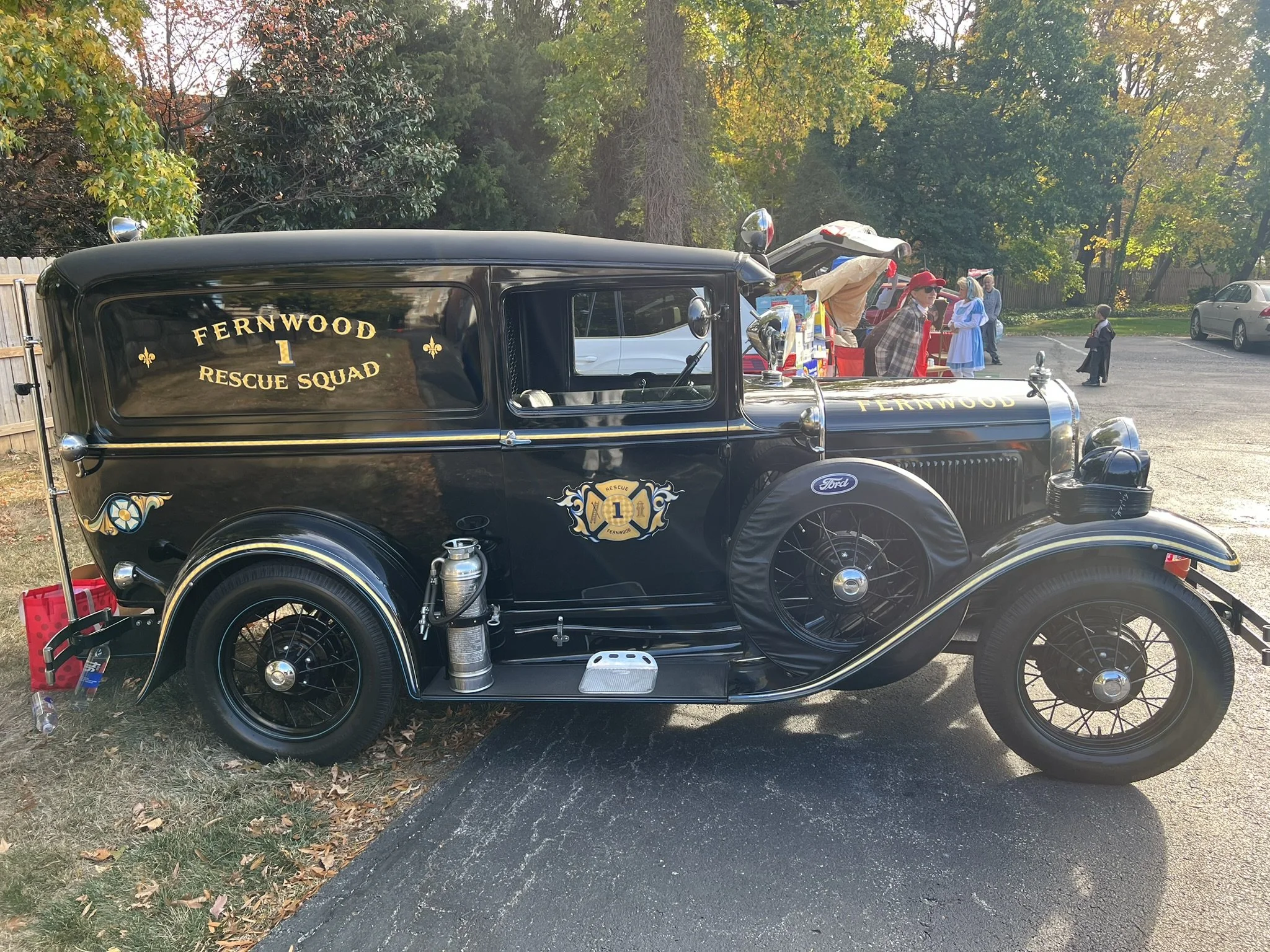 A vintage black rescue vehicle labeled 'Fernwood Rescue Squad' with gold lettering, parked outdoors on grass and pavement. In the background, there are people dressed in historical attire and a small stall with souvenirs or refreshments.