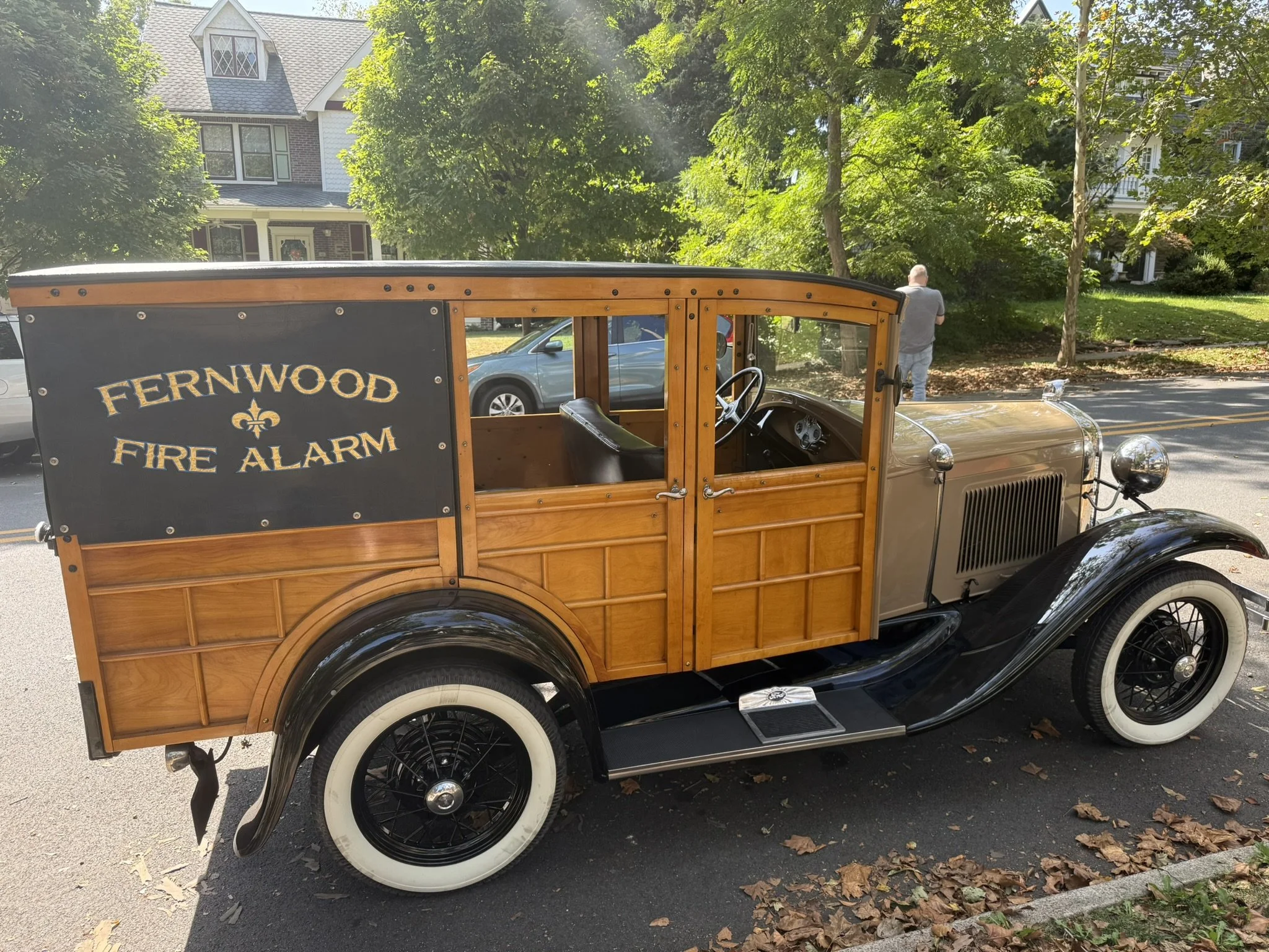 Vintage wooden fire truck with black fenders and white-walled tires parked on the street, with a sign reading 'Fernwood Fire Alarm' on the side.