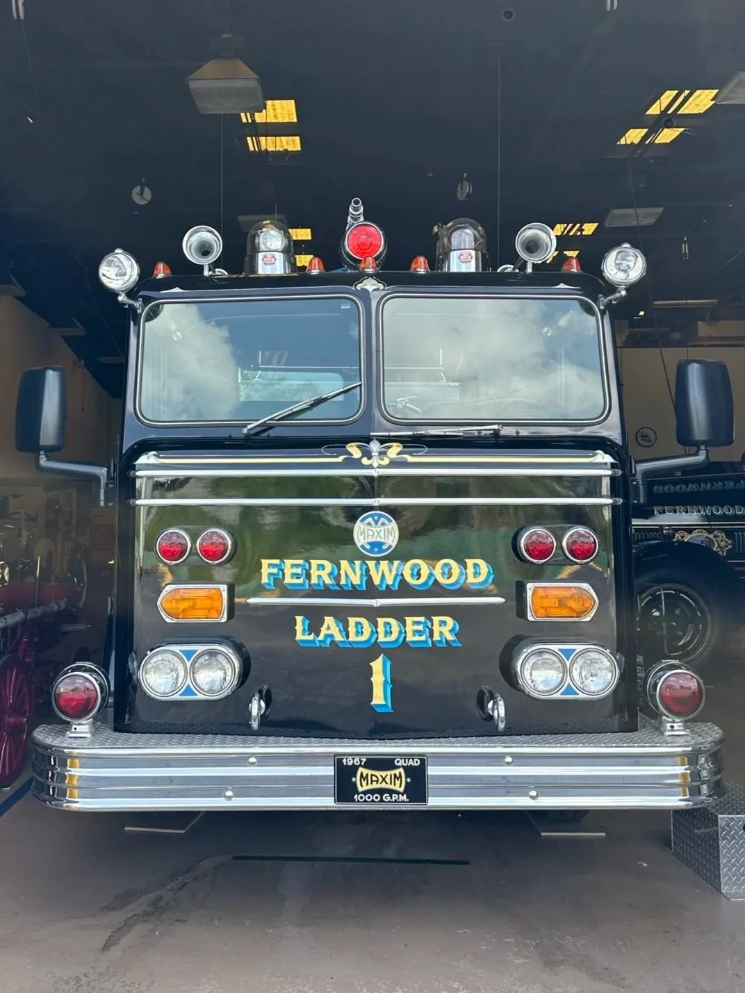 Front view of vintage fire truck labeled 'Fernwood Ladder 1' with emergency lights and sirens, inside a fire station.