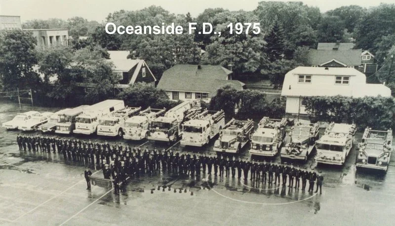 Black and white photo from 1975 showing a large group of firefighters standing in front of fire trucks at Oceanside Fire Department, with residential houses and trees in the background.