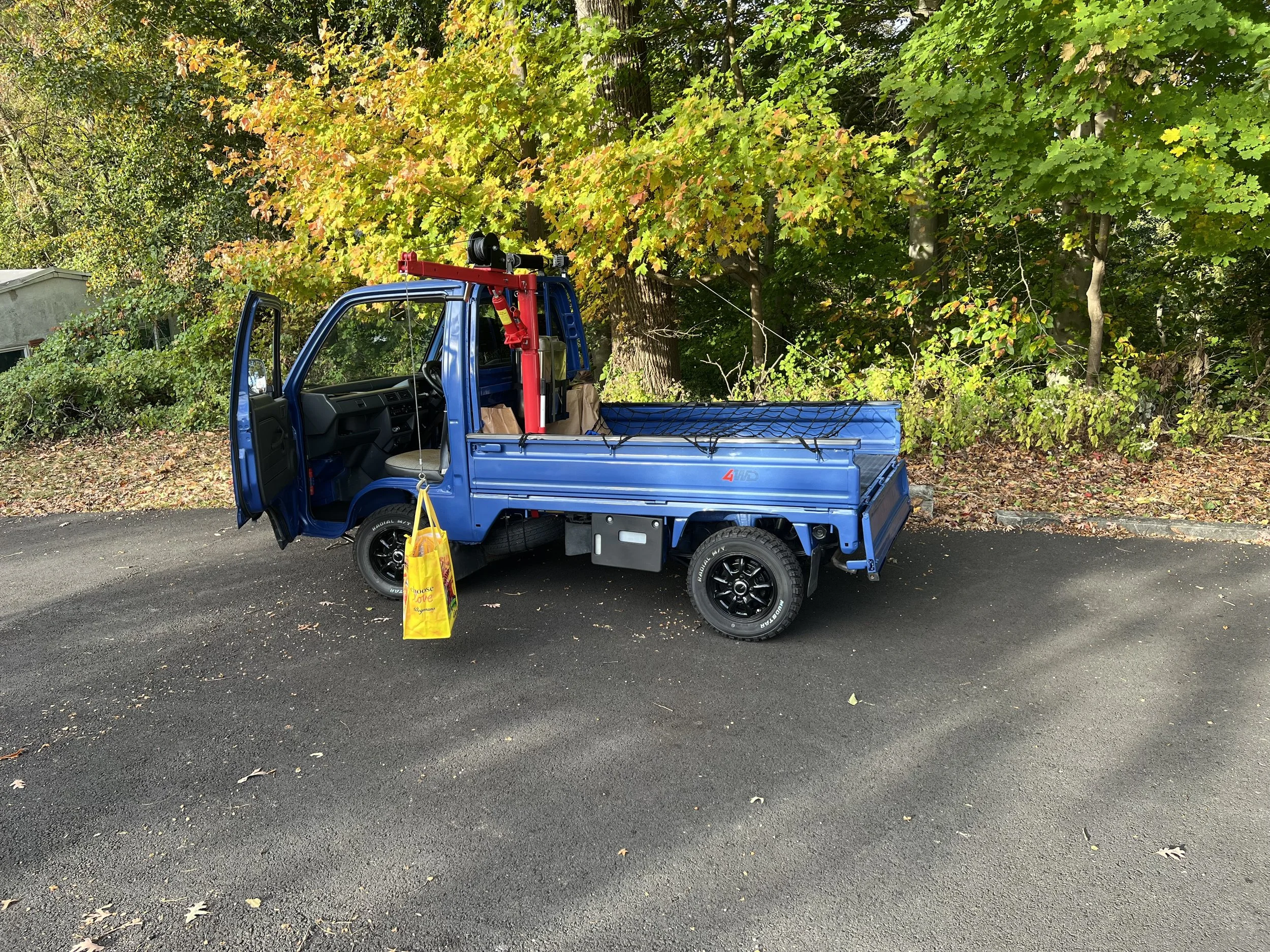 A blue mini truck parked on a dark asphalt surface with the driver’s side door open. The truck is loaded with a red tool or equipment mounted in the back and a yellow bag hanging from the side. There are trees with green and yellow leaves in the background.