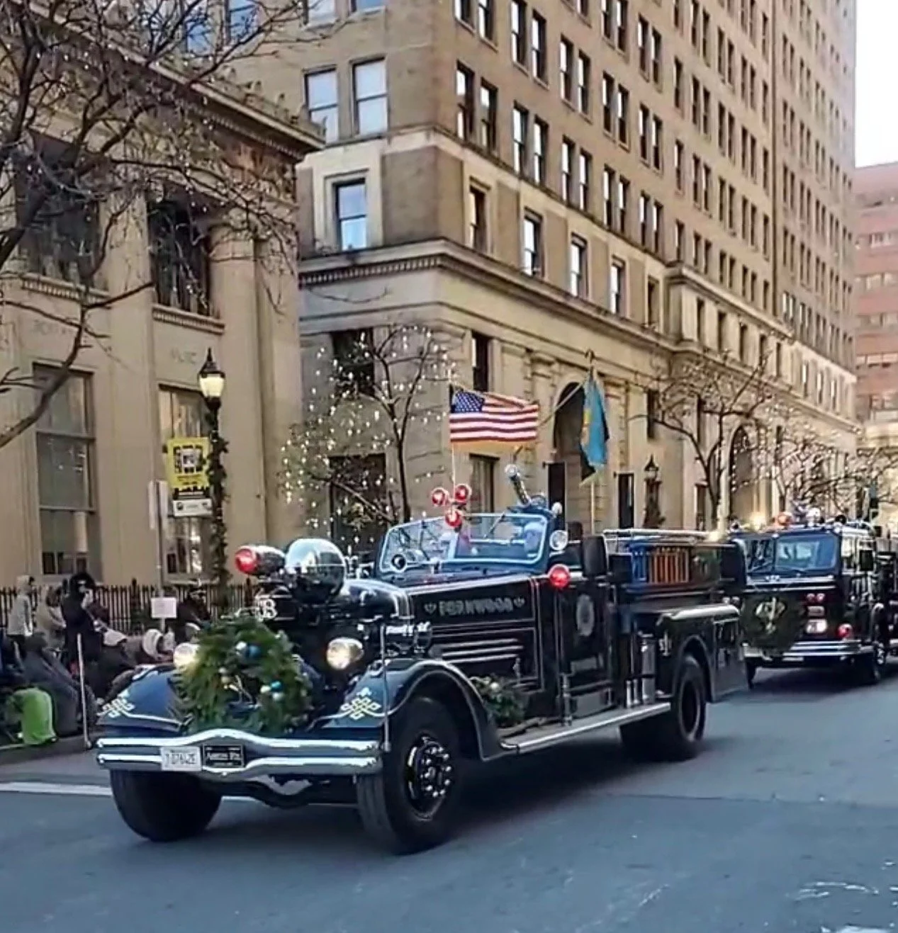 Fire truck decorated with holiday greenery and ornaments, driving on a city street with historic buildings in the background, and American and assorted flags flying.