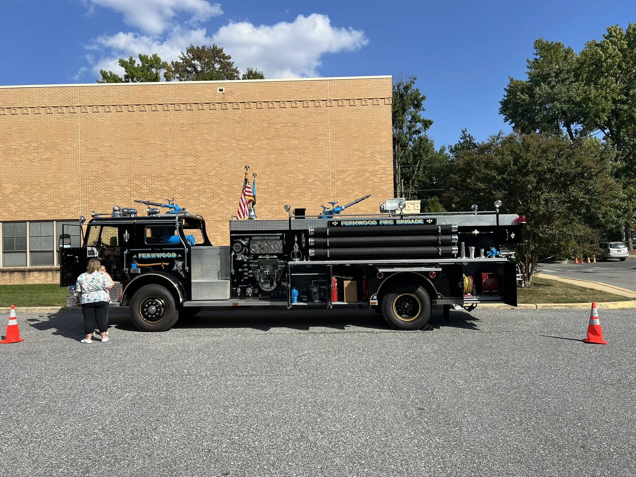 A black fire truck parked on the street with an orange cone on each side. A woman is standing next to the truck, looking at some items. There are trees, a brick building, and a blue sky with clouds in the background.