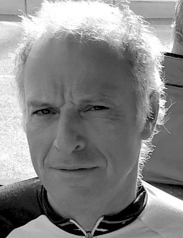 Un portrait en noir et blanc d'un homme avec des cheveux grisonnants, portant une veste de sport et la mer en arrière-plan.