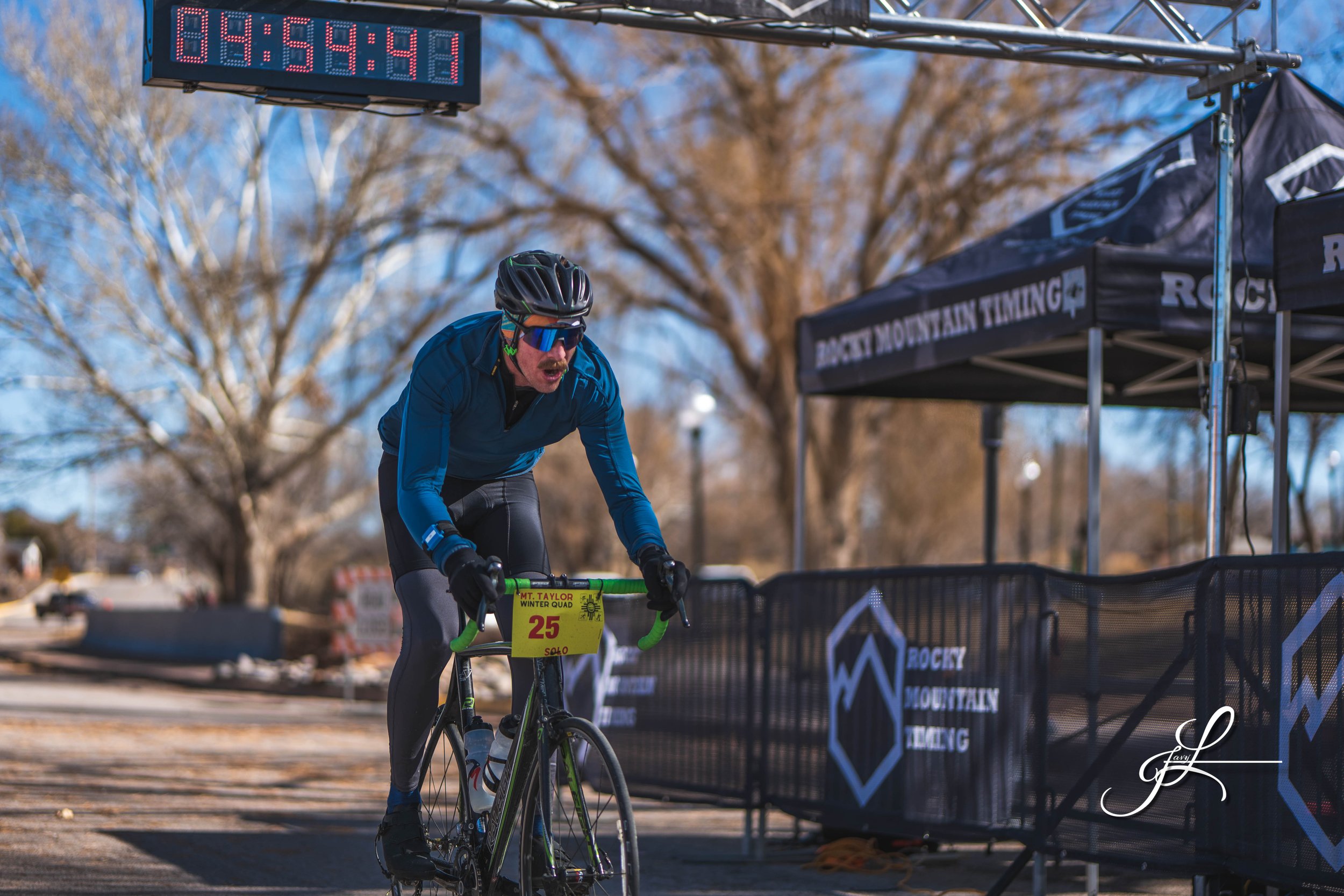 Male cyclist crossing the finish line during a race with a digital timer overhead showing 94:54:41, wearing a helmet, sunglasses, and sports gear, with a race number 25 on his bike, under a tent labeled 'Rockey Mountain Tering' in a park with leafless trees.