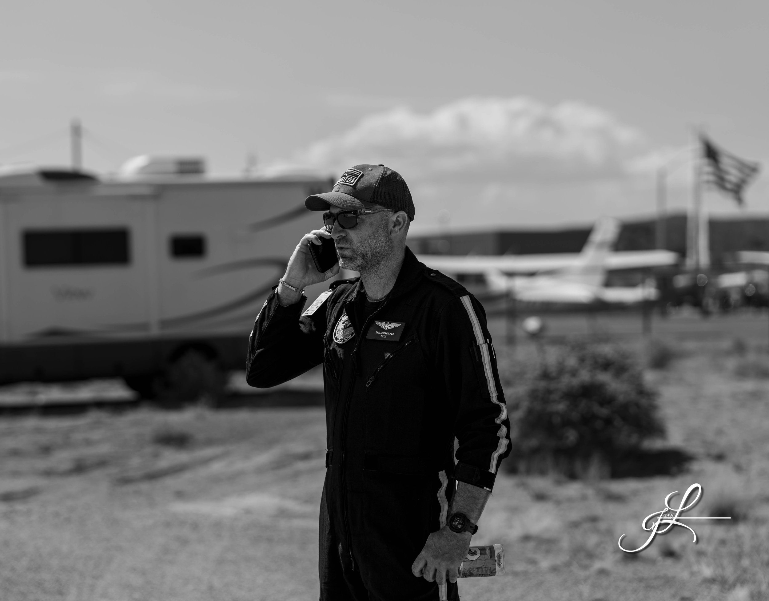 A man in a pilot uniform talking on a cellphone outdoors, with an RV, airplanes, and an American flag in the background.