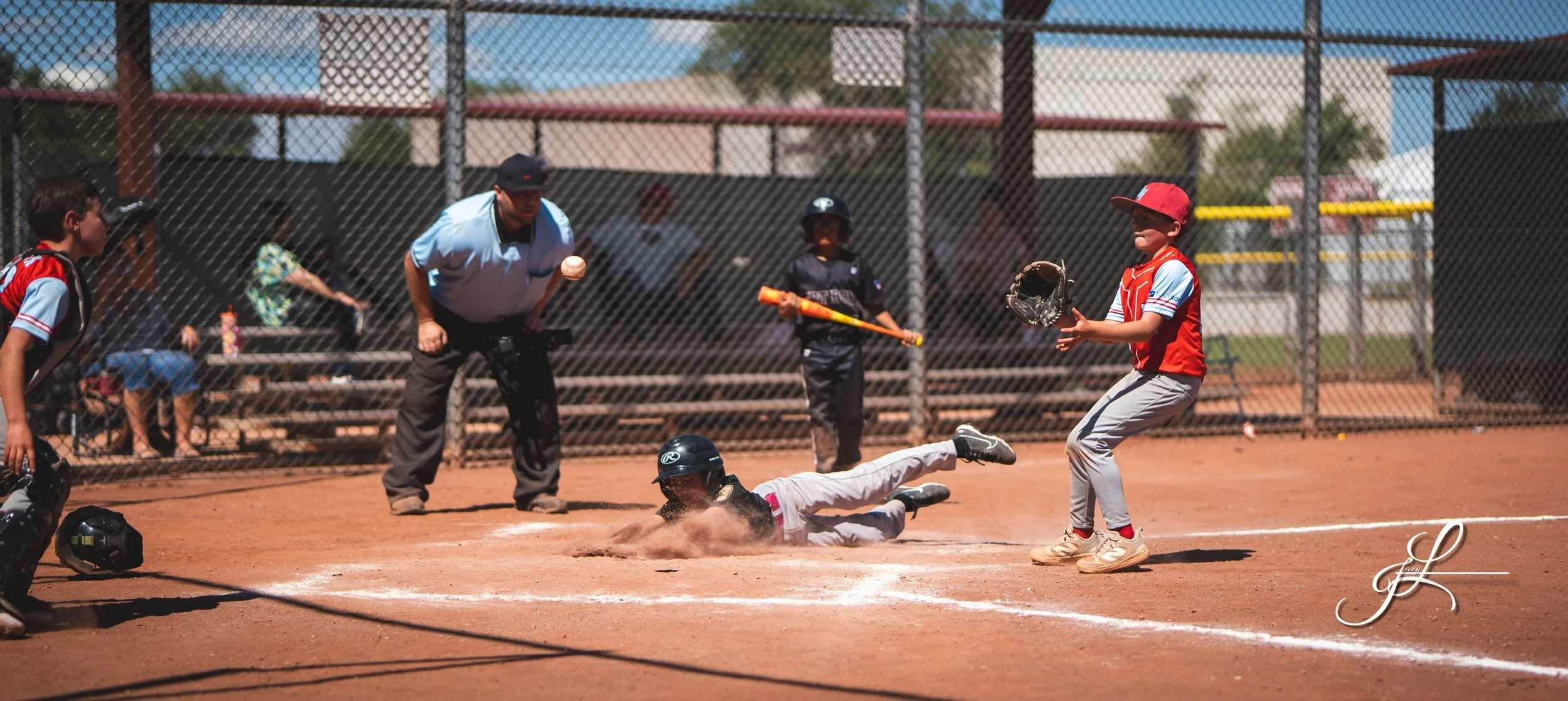 A youth baseball game with a player sliding into home plate as the catcher catches the ball, an umpire observes, and other players stand nearby on a dirt field, with a chain-link fence and bleachers in the background.