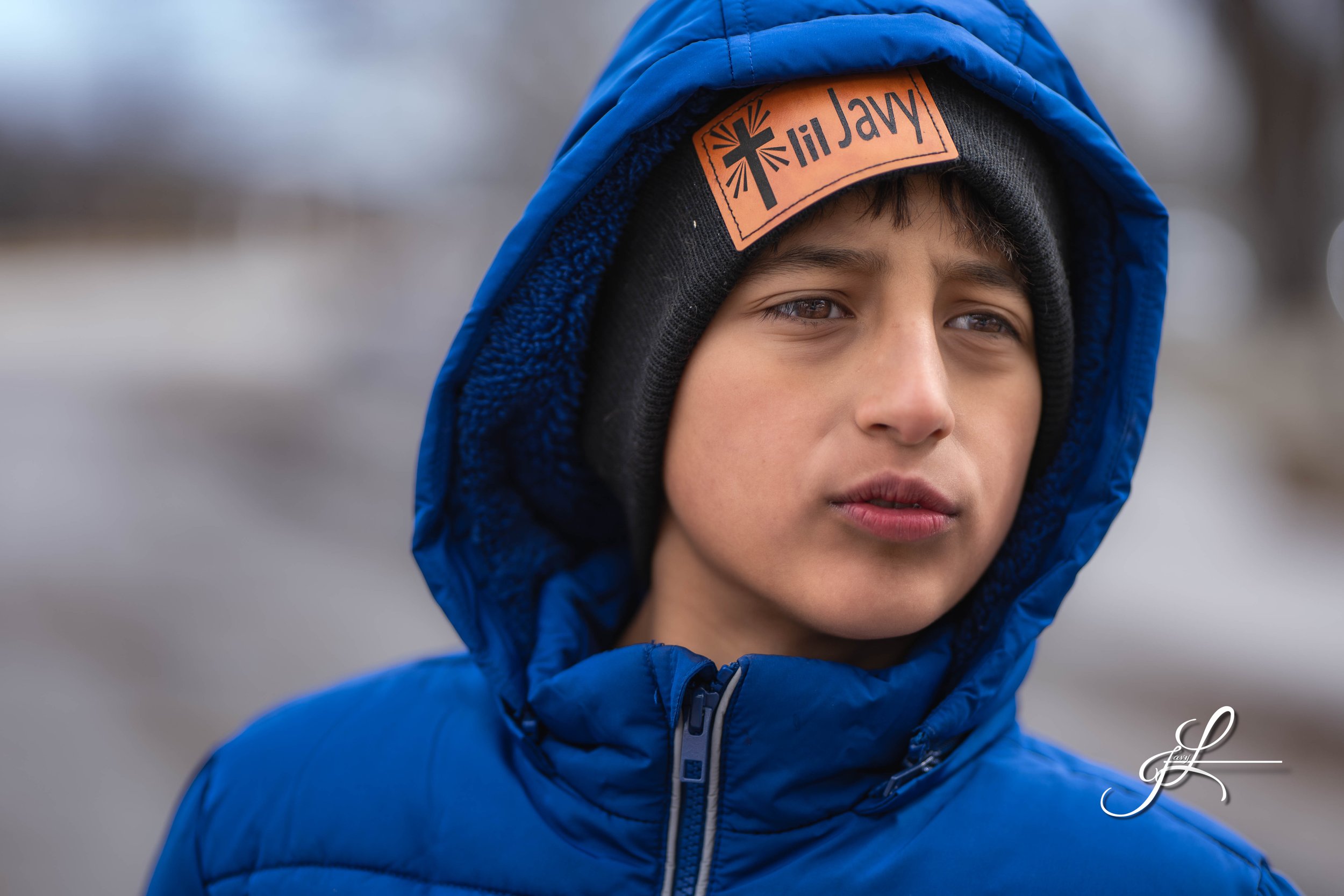 Close-up of a boy wearing a blue winter jacket and a black hat with a logo, outdoors in a cold environment.