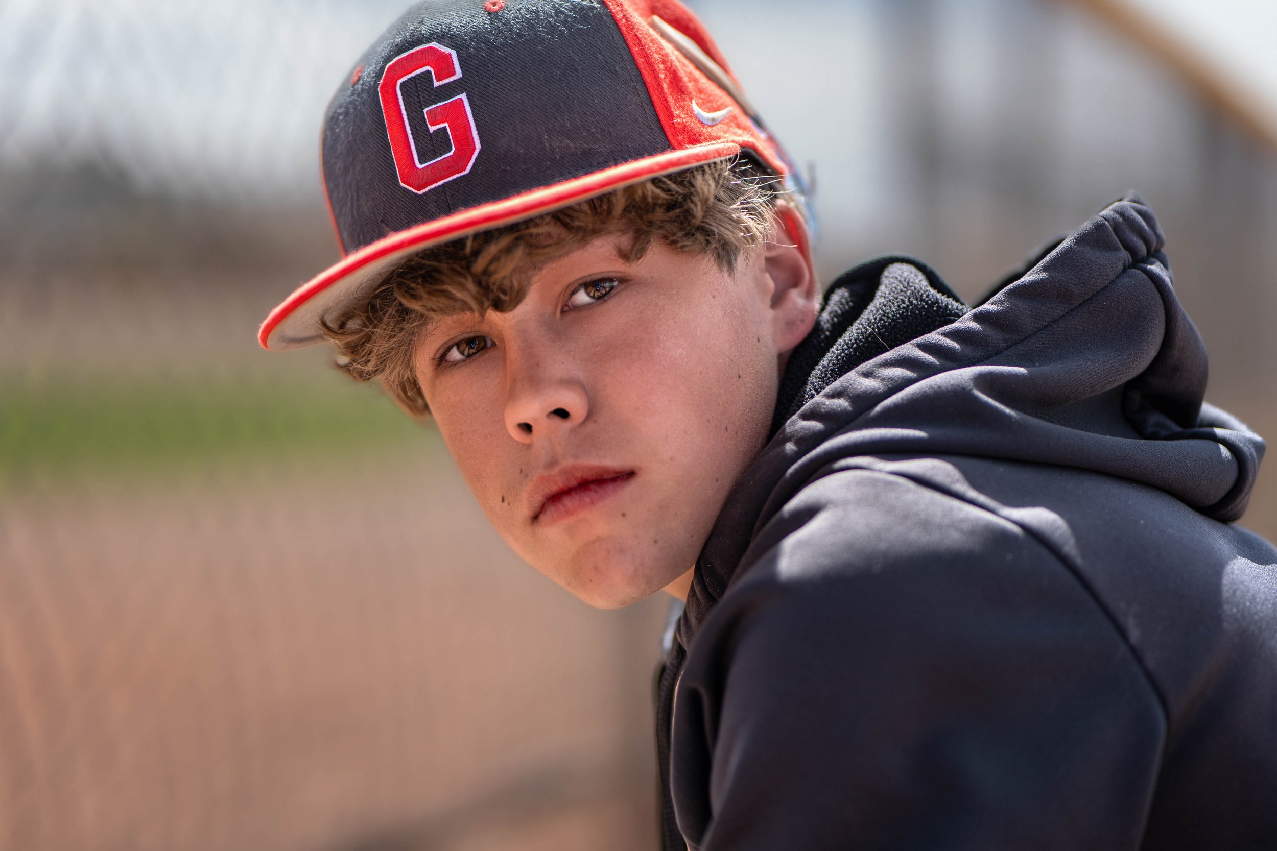 A teenage boy with light skin and curly brown hair wearing a black and red baseball cap with a large red letter G, a black jacket, and a gray hoodie, outdoors on a sunny day with a blurred background.