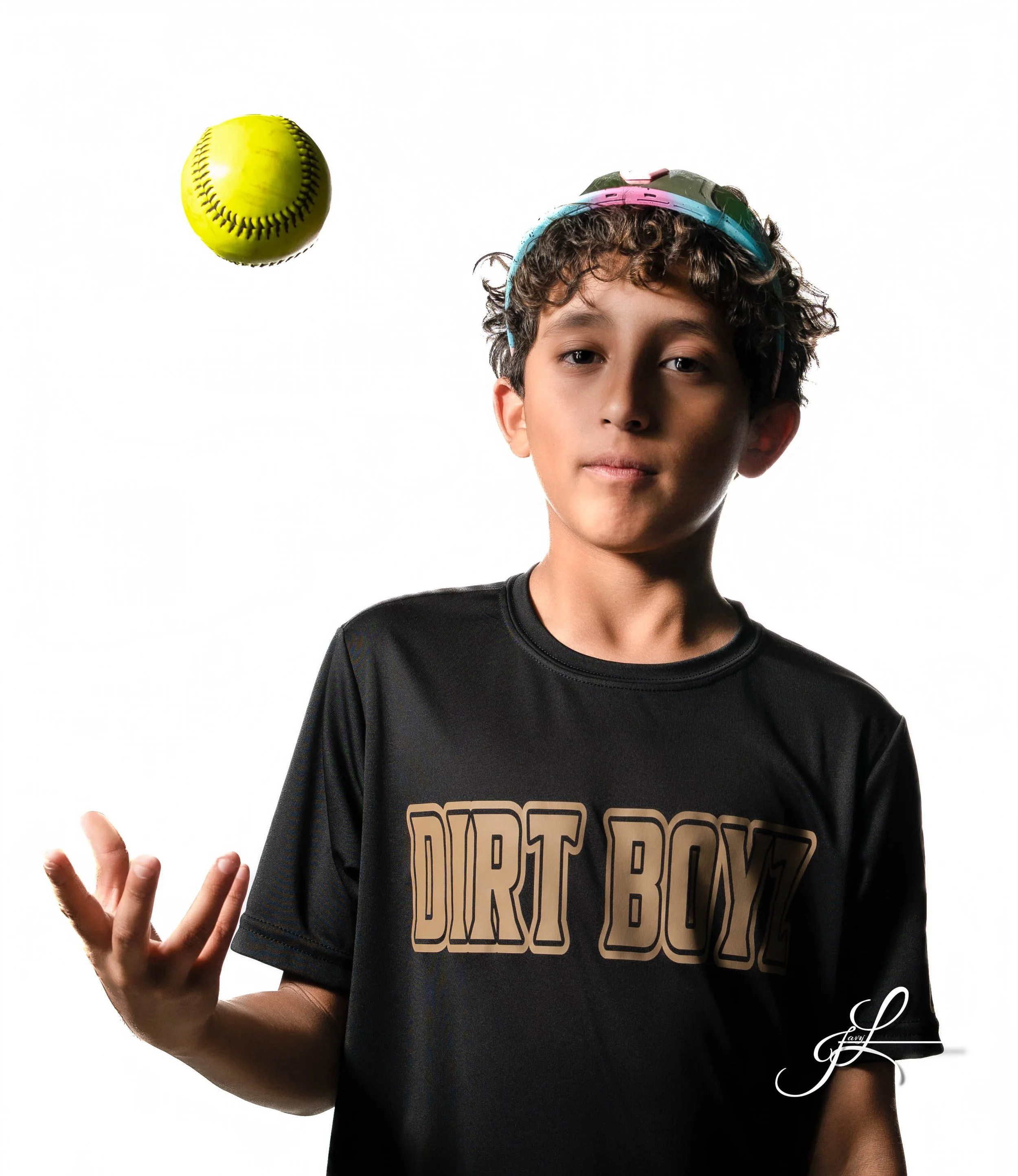 A young boy with curly hair wearing a black t-shirt that says 'DIRT BOYZ' and a colorful pair of sunglasses on his head, standing against a white background, with a yellow softball floating in the air nearby.