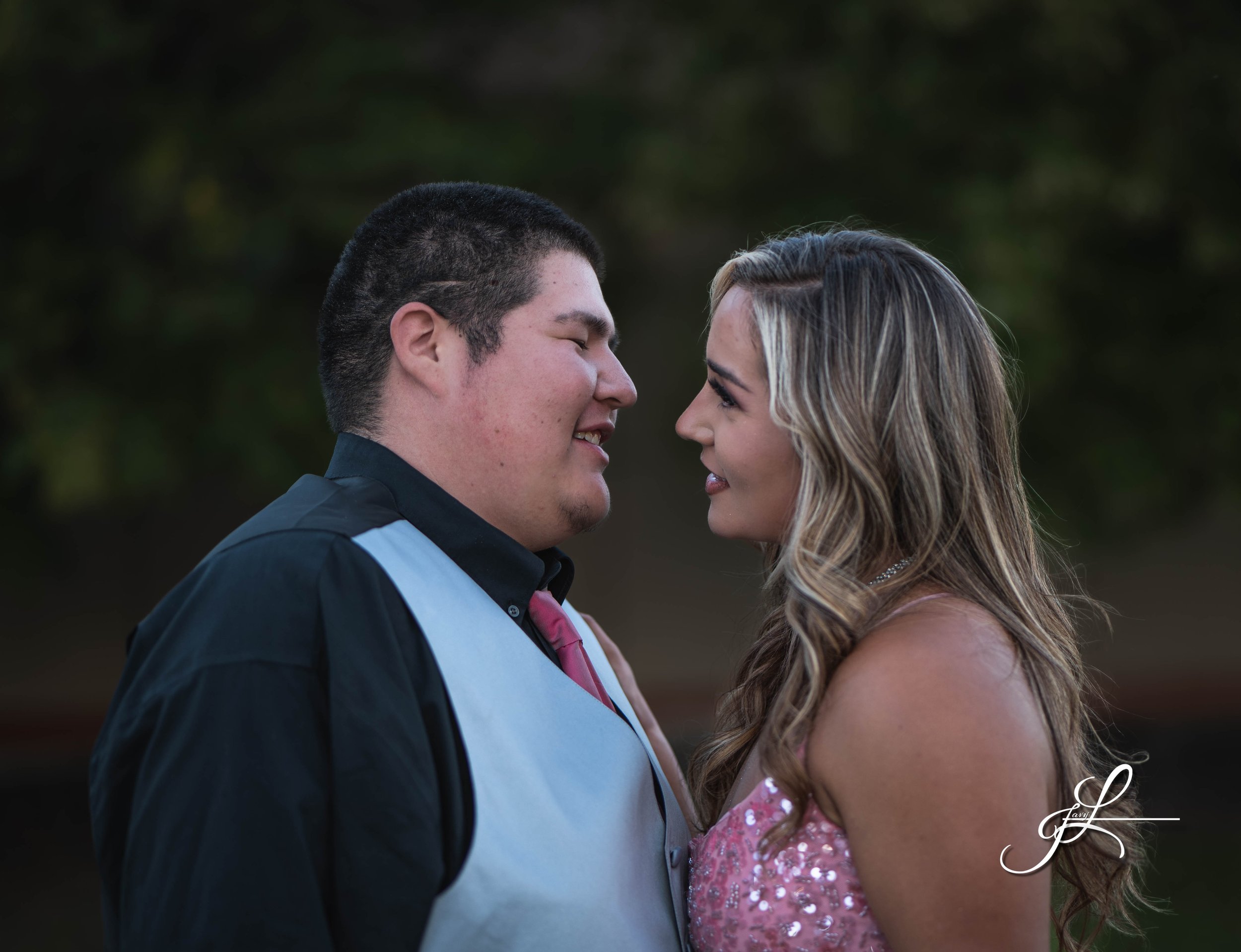 A young man and woman face each other closely, smiling with their noses nearly touching, outdoors with a blurred green background.