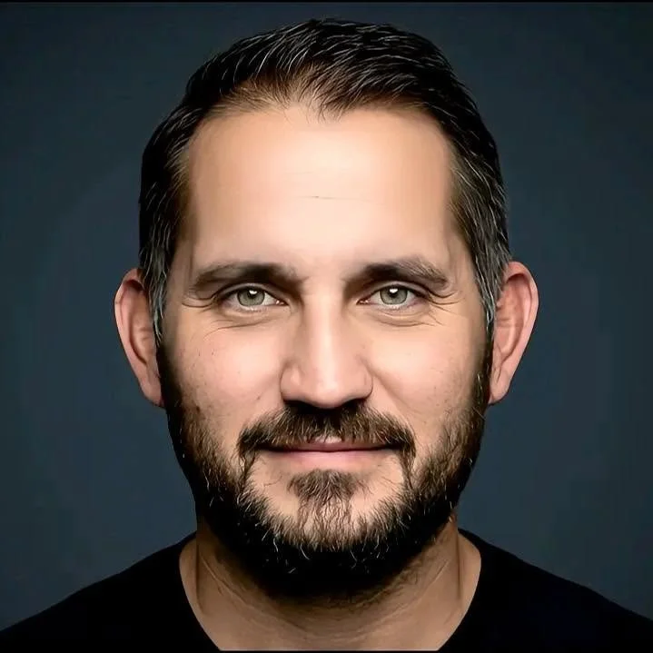 Close-up portrait of a man with dark hair, a full beard, and blue eyes, smiling slightly against a dark background.