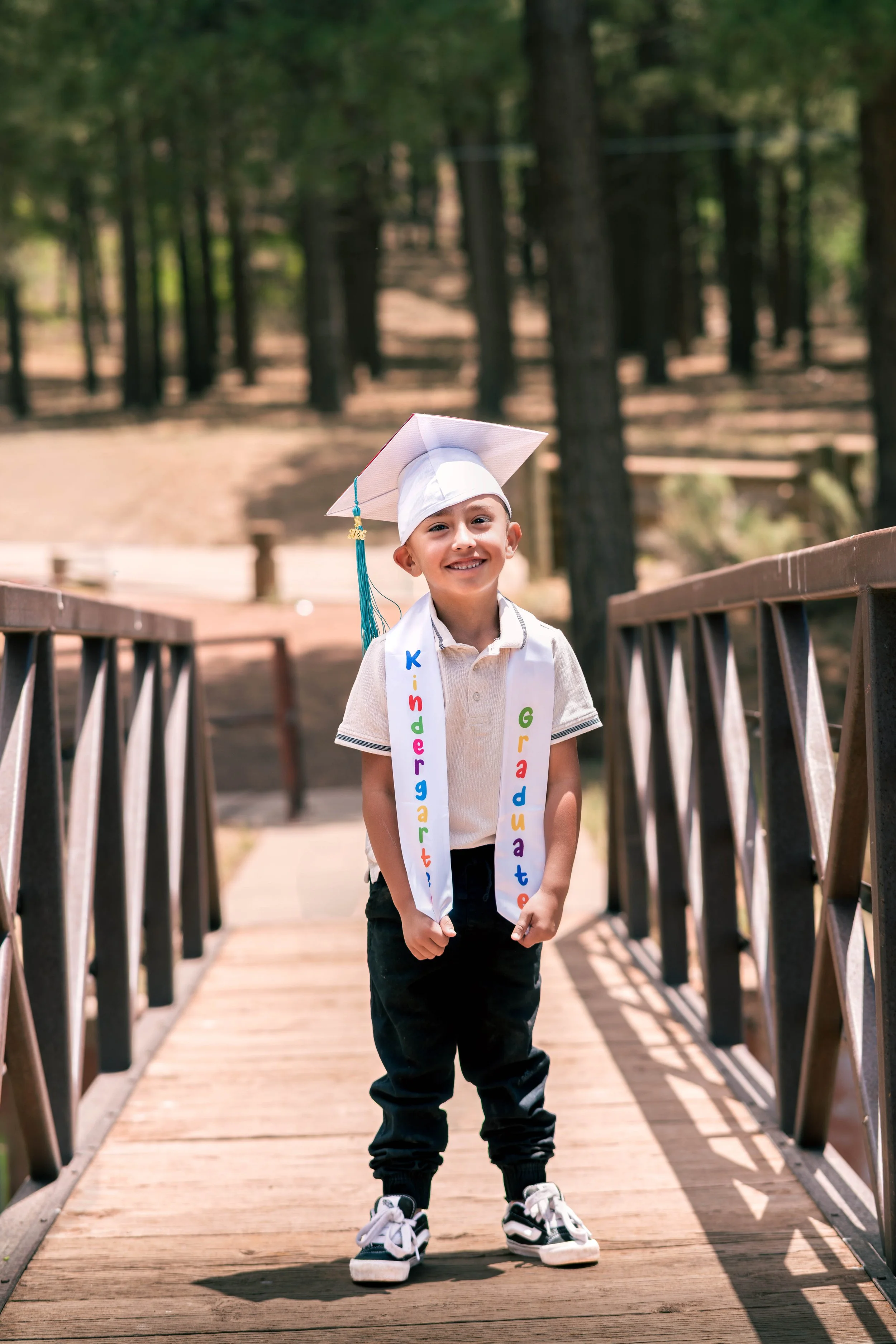 A young boy standing on a wooden bridge in a forest, wearing a white graduation cap and sash with colorful letters, black pants, and sneakers, smiling at the camera.