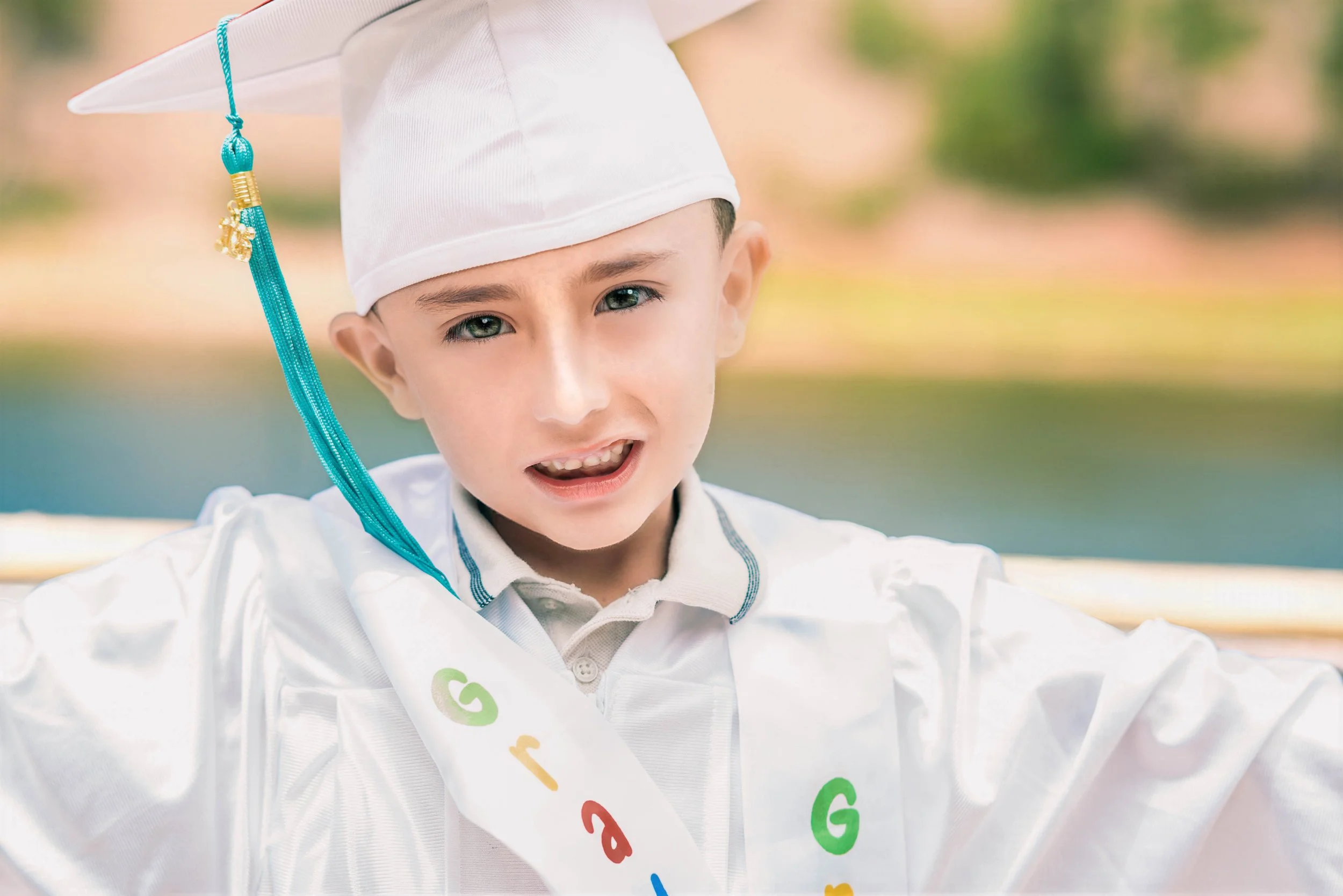 Young boy wearing a graduation cap and gown with a colorful alphabet sash, smiling outdoors.