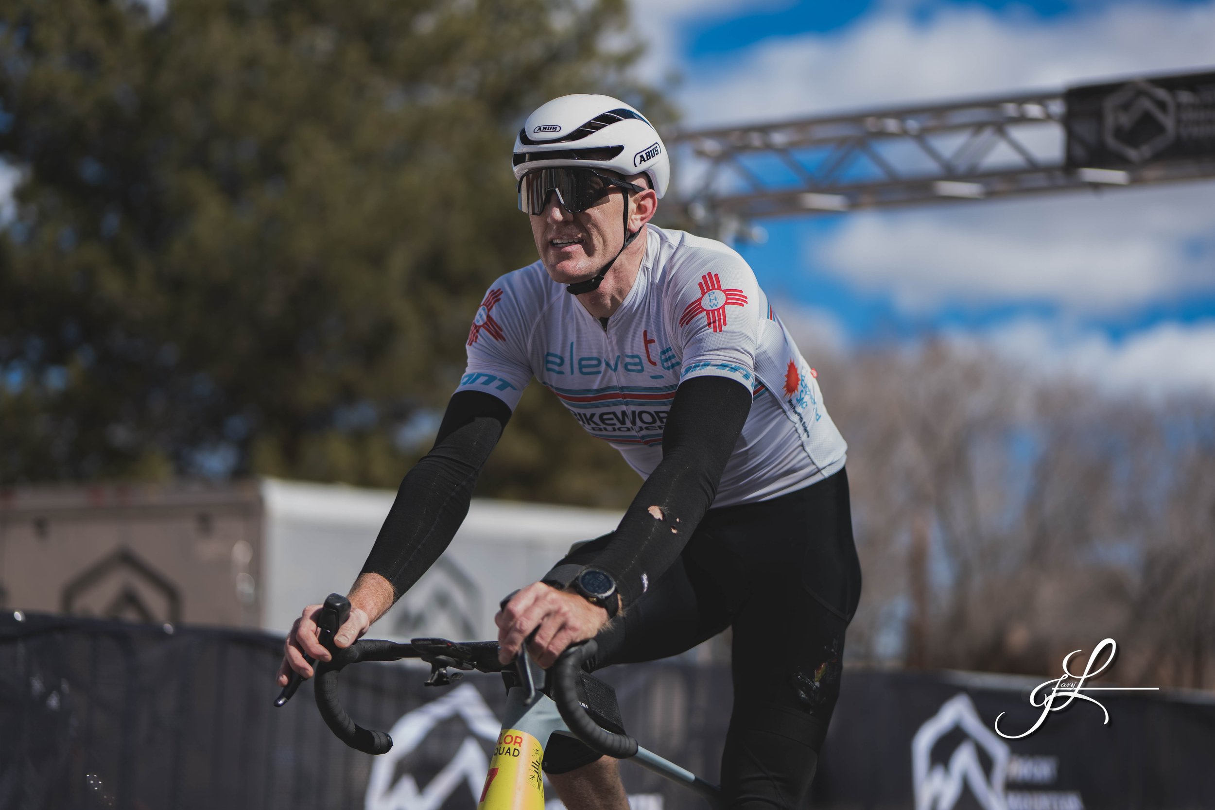 A man riding a bicycle outdoors during daytime, wearing a white helmet, sunglasses, a white cycling jersey with various sponsor logos, and black arm warmers. The background shows trees and a blue sky with some clouds.
