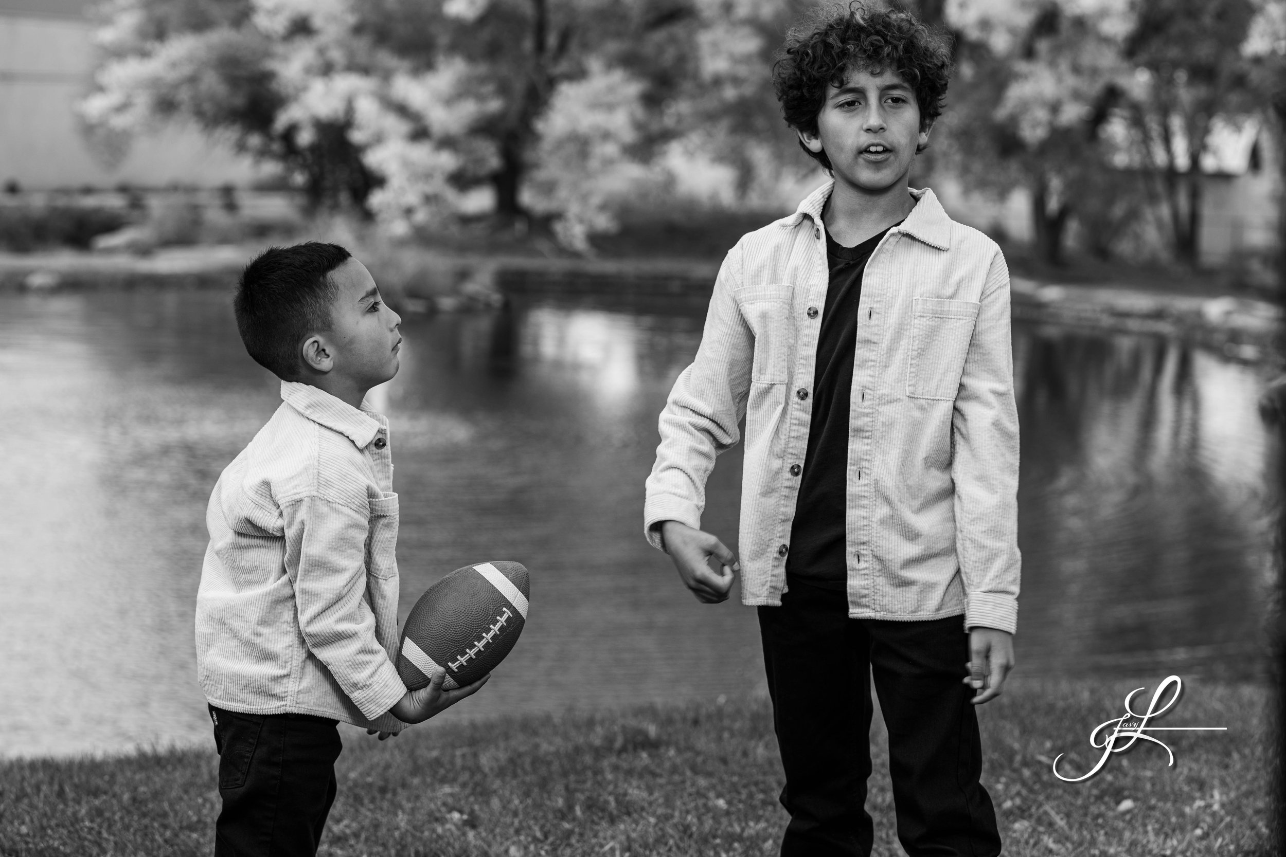 Two boys outdoors by a river, one holding a football, the other talking, with trees in the background