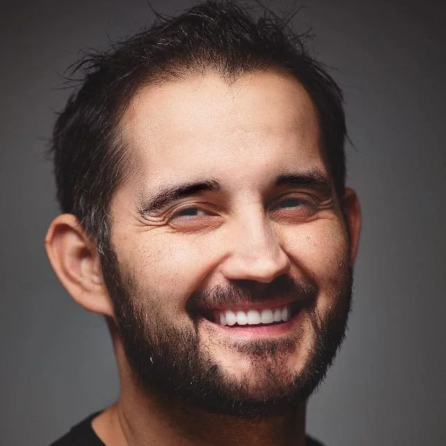 Close-up portrait of a smiling man with dark hair and beard, looking at the camera against a plain gray background.