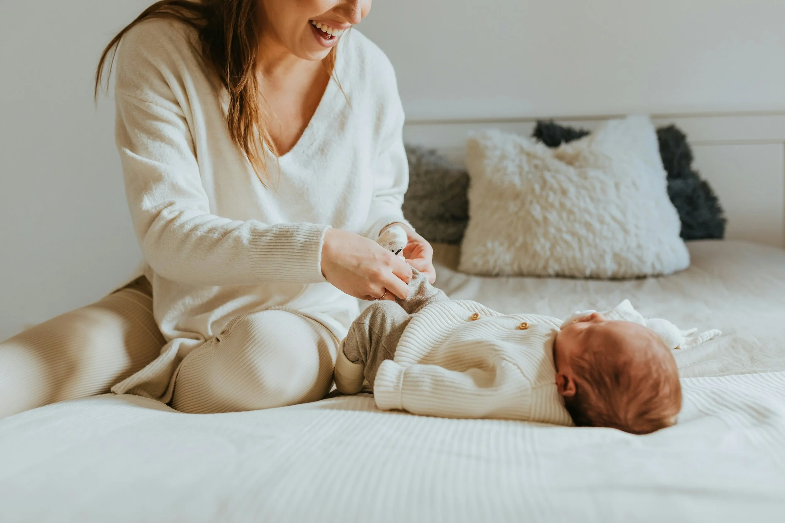 A woman playing with a baby on a bed, smiling and dressed in cozy clothing, with soft pillows in the background.