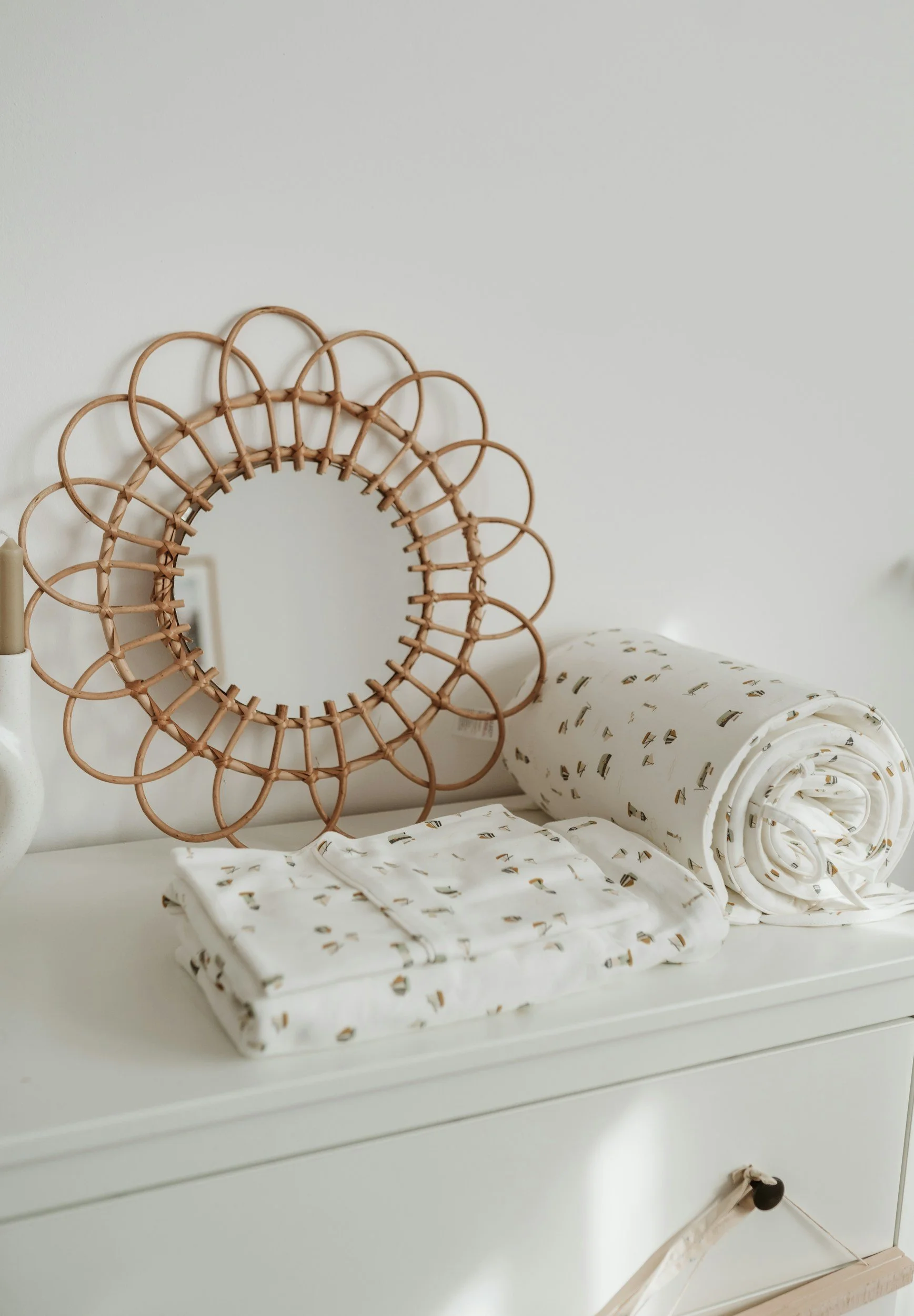 Decorative rattan mirror and folded patterned fabric on a white dresser.
