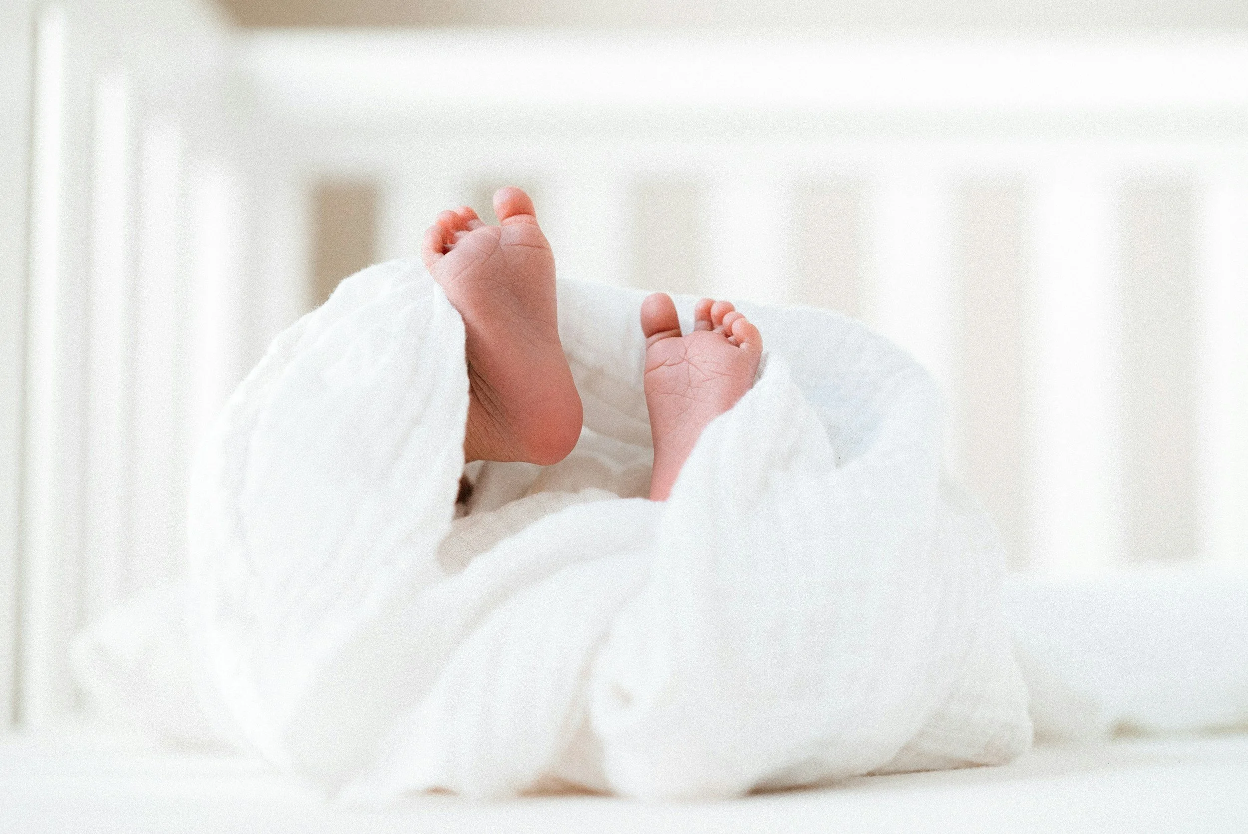 Close-up of a newborn baby lying on a white blanket with tiny feet and toes, in a bright nursery setting.
