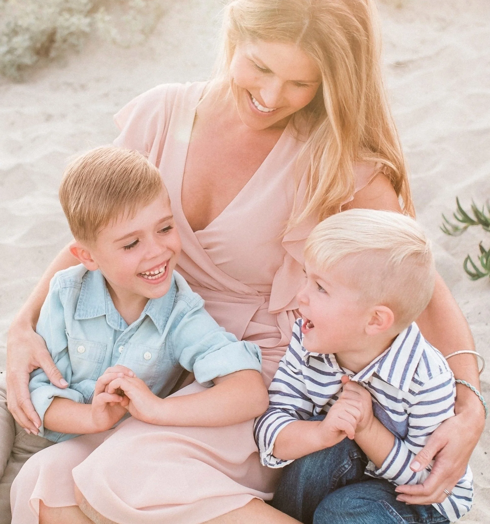 A woman with long red hair smiling while sitting on the sand with two young boys, one with blonde hair and one with light brown hair, both smiling and engaging with each other.