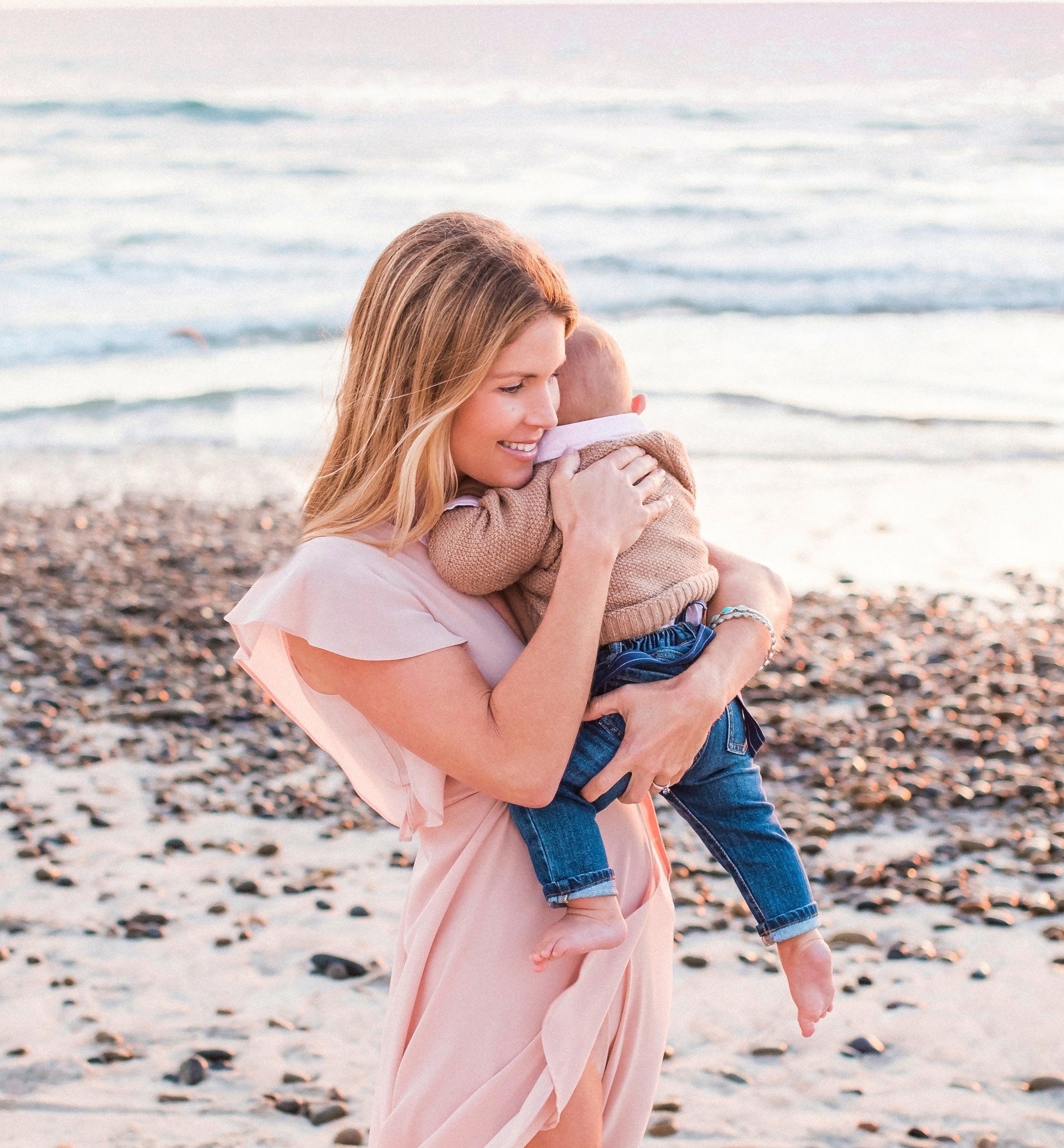 A woman holding a young child at the beach near the water, smiling, during sunset.