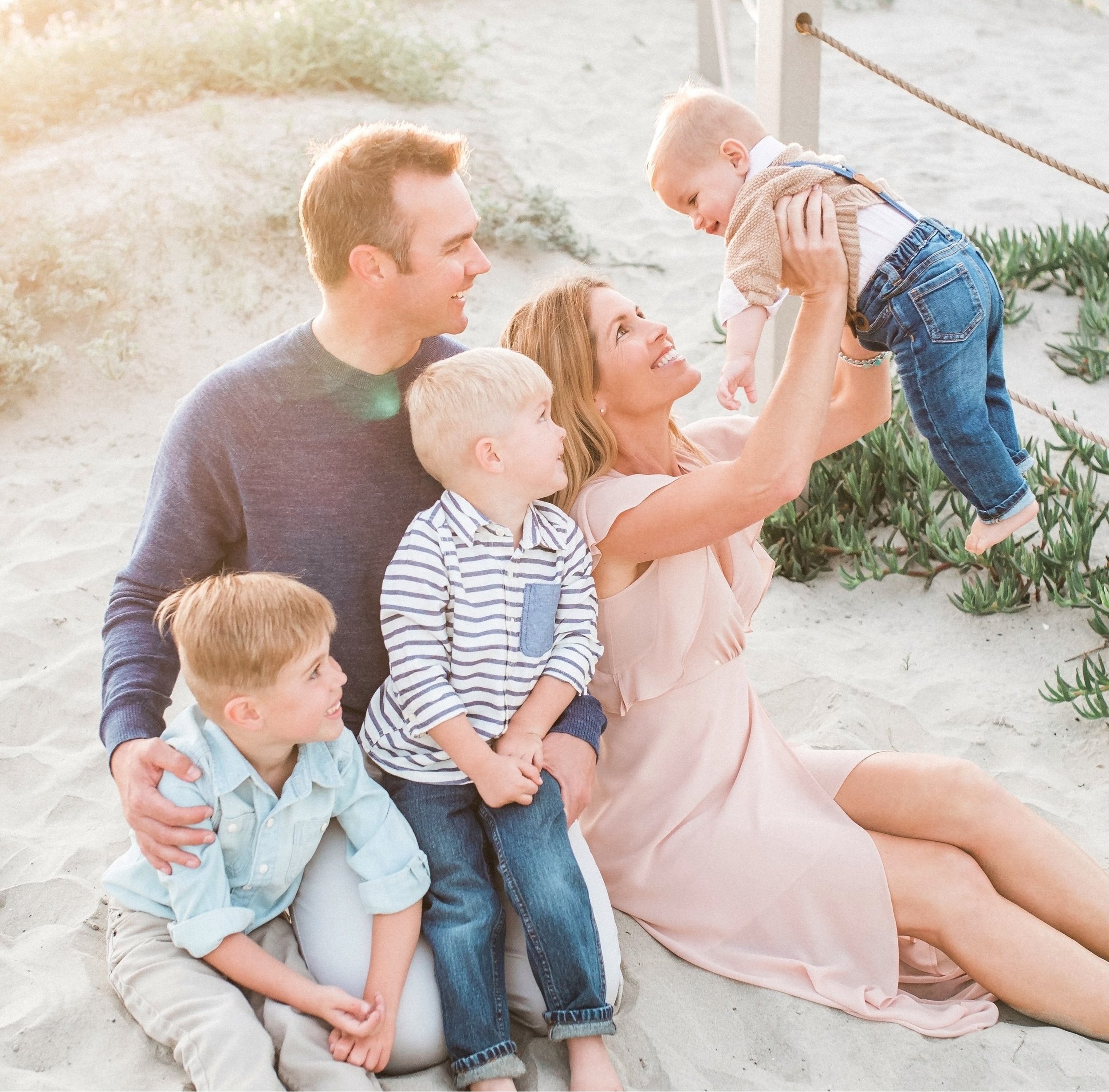 Family of five sitting on the beach, smiling, with a woman lifting a young child in the air, during sunset.