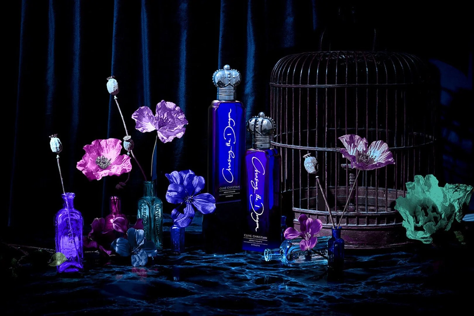 Assorted colored perfume bottles and flowers arranged on a dark blue surface with a dark curtain background, with an empty birdcage on the right.