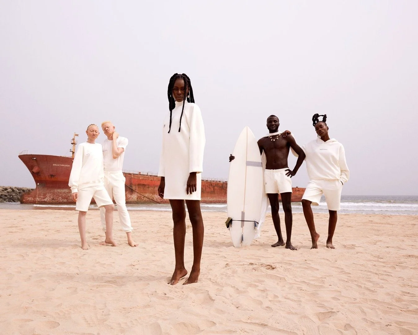 A diverse group of five models stands on a sandy beach with an abandoned ship in the background. They are dressed in white clothing, with one guy holding a surfboard. The atmosphere is calm and the sky is overcast.