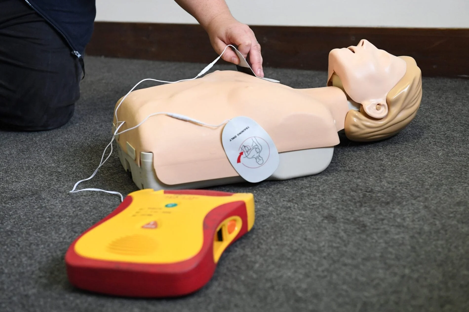Hands performing CPR on a training dummy lying on a gray carpeted floor with an automated external defibrillator (AED) nearby.