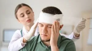 A woman in medical gloves applying a treatment to a man's head, who has a bandage wrapped around his forehead, in a clinical setting.