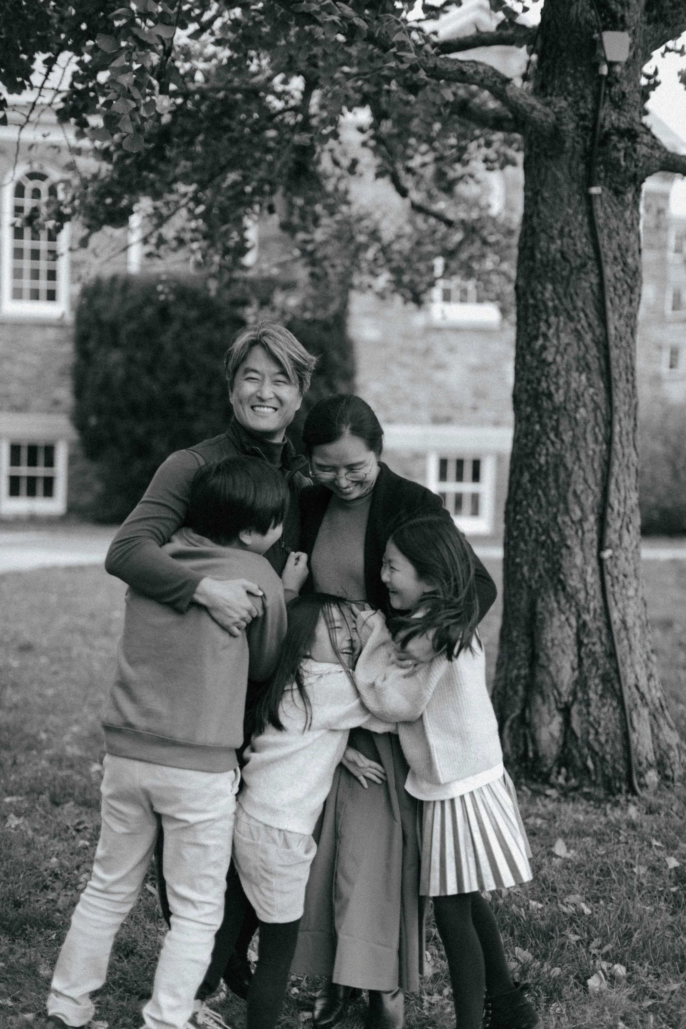 A group of five people, including children and adults, hugging and smiling outdoors near a large tree and a brick building, captured in black and white.