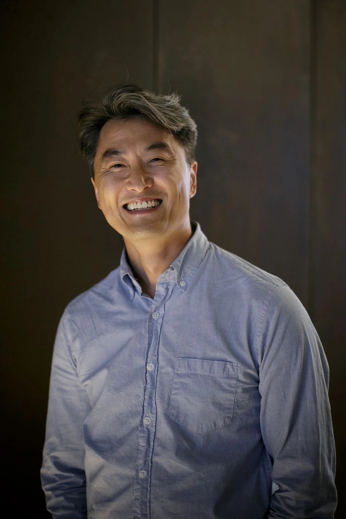 A man smiling and winking at the camera, wearing a light blue button-up shirt, with a dark background.