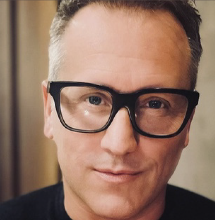 Close-up of a man wearing black-framed glasses, smiling softly, with short brown hair and fair skin.