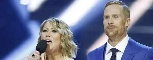 A woman speaking into a microphone and a man standing beside her, both on a stage with a blue background.