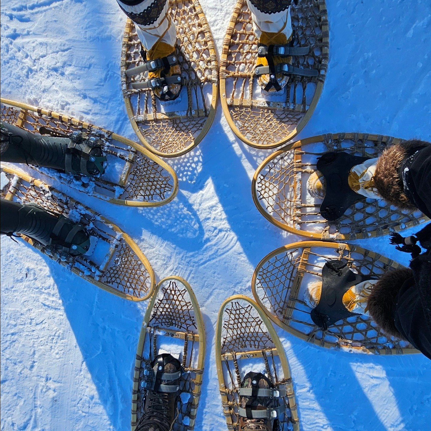 Six snowshoes arranged in a circle on snow, with pairs of snow boots inside each, and a camera on one of the snowshoes. The photo is taken from above during daylight.