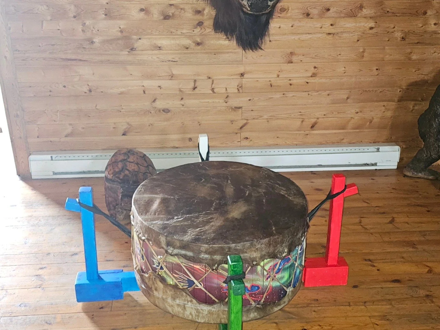 An indoor scene featuring a small drum with a colorful design, secured to four small wooden stands painted red, blue, green, and white. The background has wooden paneling along a wall, with a baseboard heater visible below.