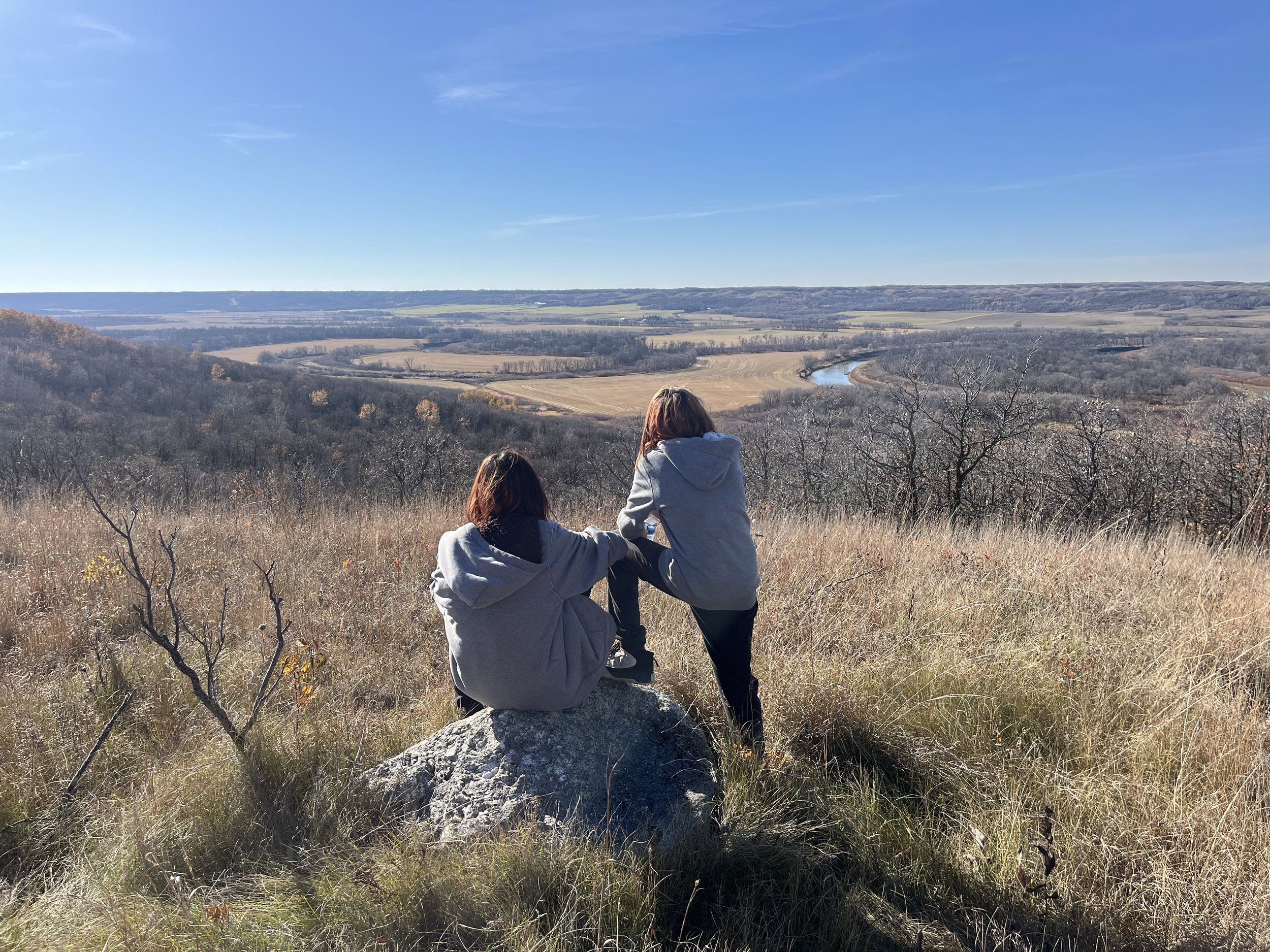 Two women in gray hoodies sitting and standing on a large rock, overlooking a scenic landscape of fields, trees, and a river under a blue sky.