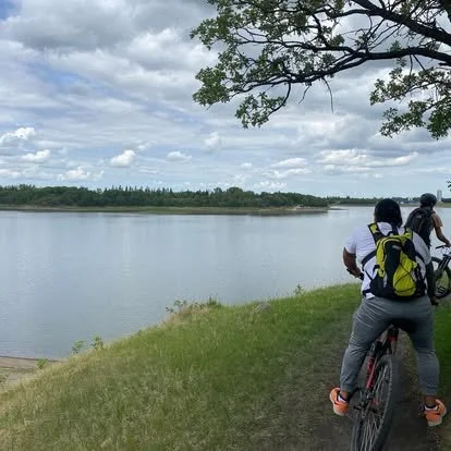 Two people riding bicycles along a grassy riverbank under a cloudy sky, with a water body and trees in the background.