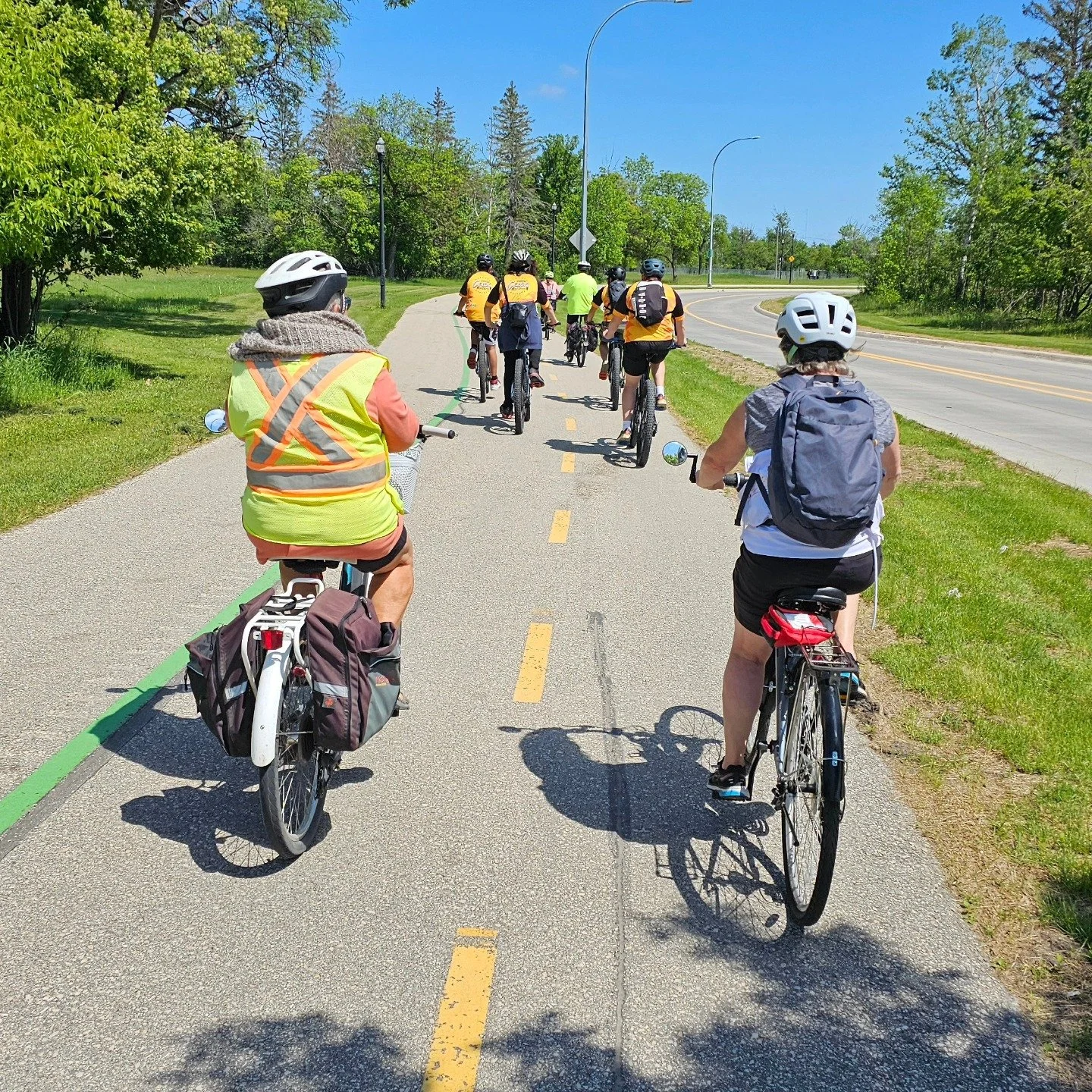A group of people riding bicycles on a dedicated bike path next to a road, surrounded by green trees and a bright blue sky.