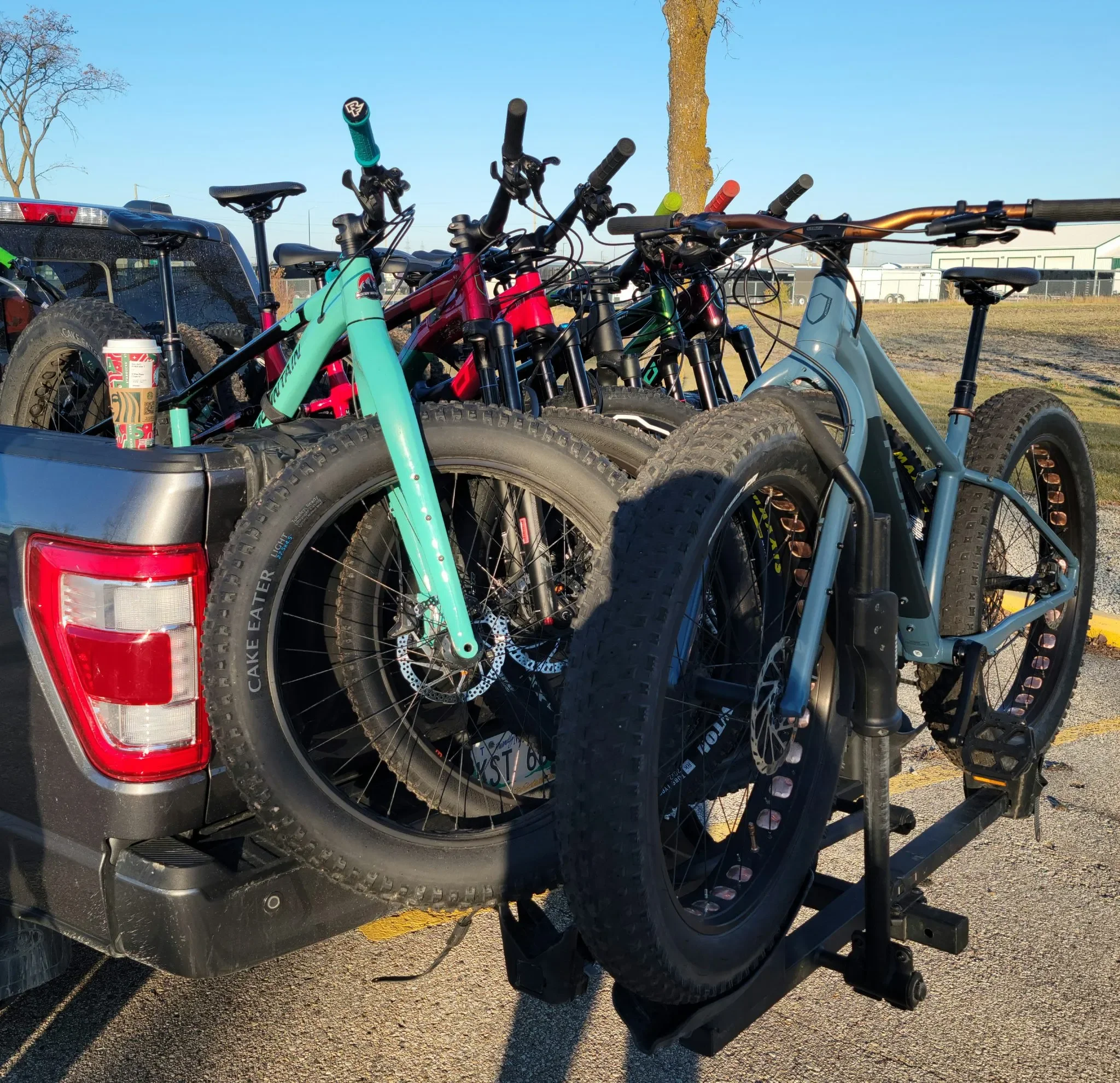 Pickup truck bed loaded with several mountain bikes, some with colorful frames, and a travel mug, in a parking lot with a tree and industrial buildings in the background.
