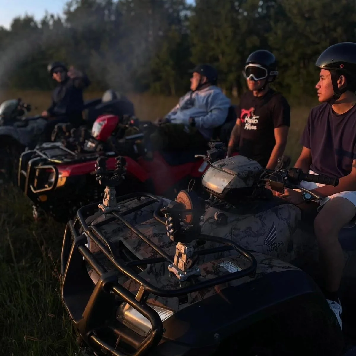 Four men sitting on all-terrain vehicles (ATVs) in a grassy field during dusk, with a forest in the background.