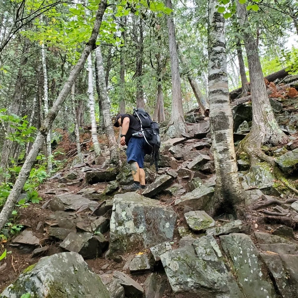 A person with a backpack hiking on a rocky trail through a wooded forest.