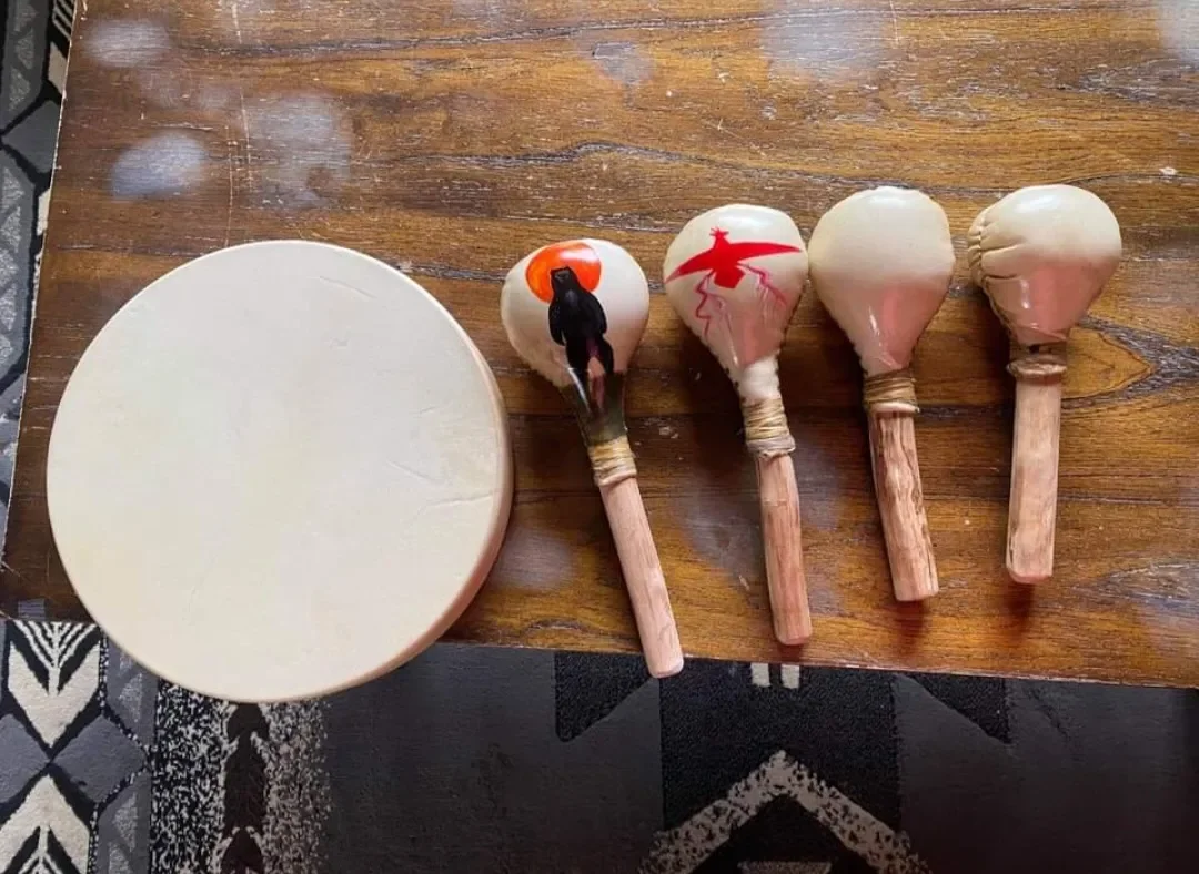 Four wooden maracas with painted designs and a round white drum on a wooden table.