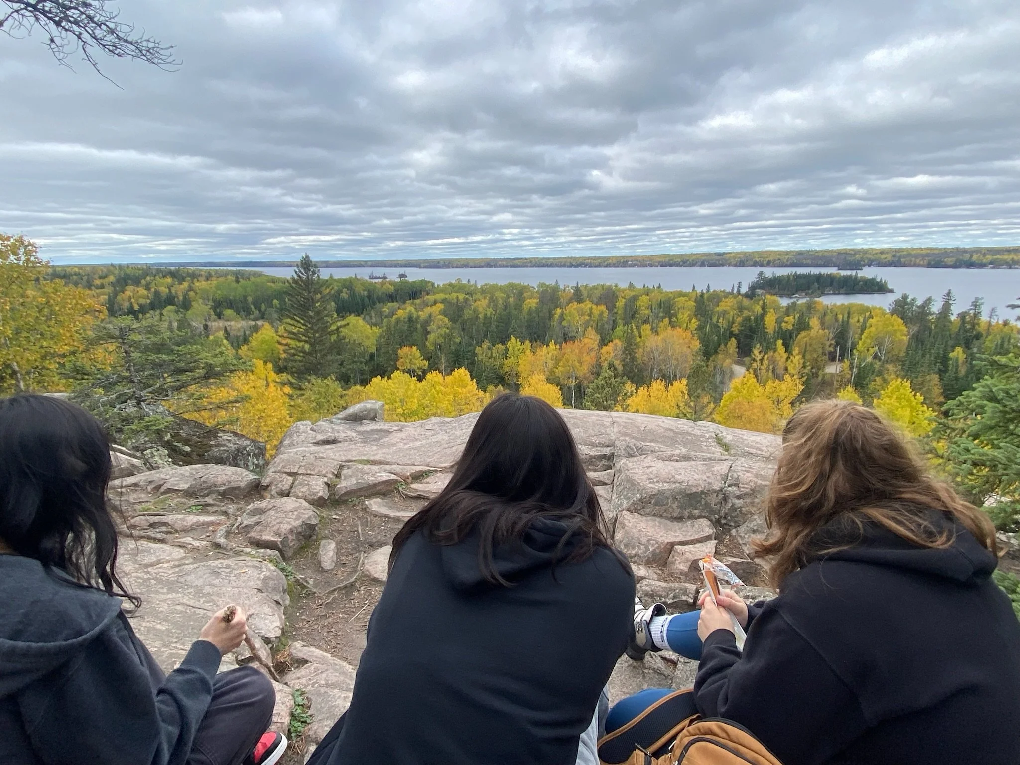 Three women sitting on rocks on a hilltop, overlooking a forested area with yellow and green trees and a large body of water under a cloudy sky.
